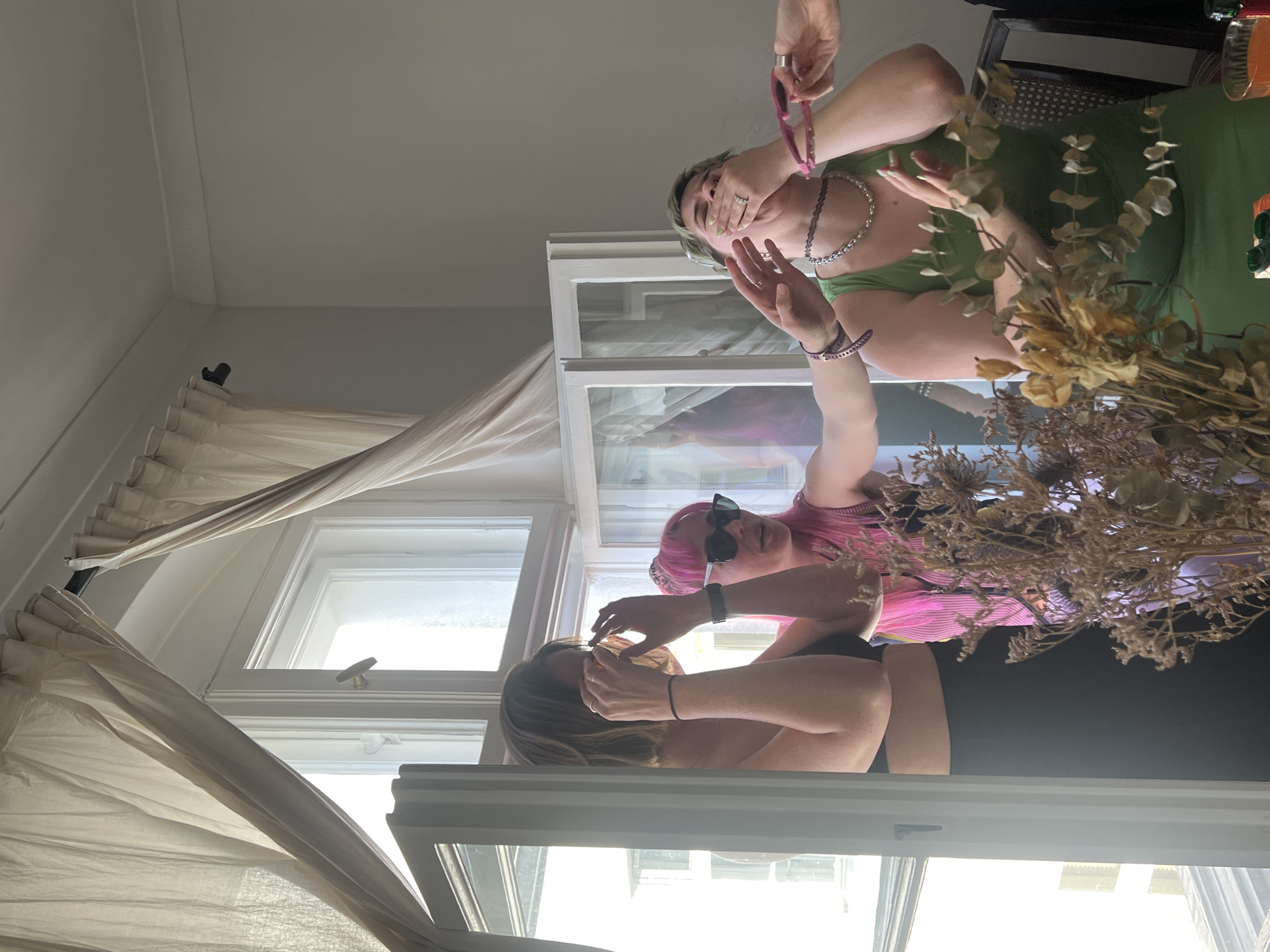 Three individuals are inside a room with natural light. The first individual, wearing a green top and silver necklace, is covering their mouth with their hand. An arm with a pink glasses cord is visible above them. The second individual, wearing a pink shirt and sunglasses, has an arm extended above the first individual. The third individual, wearing a beige top, is holding their face with their hand. Dried flowers are in the middle of the scene. Curtains are visible by the window. The location is not identifiable as Berlin, Germany from the image.
