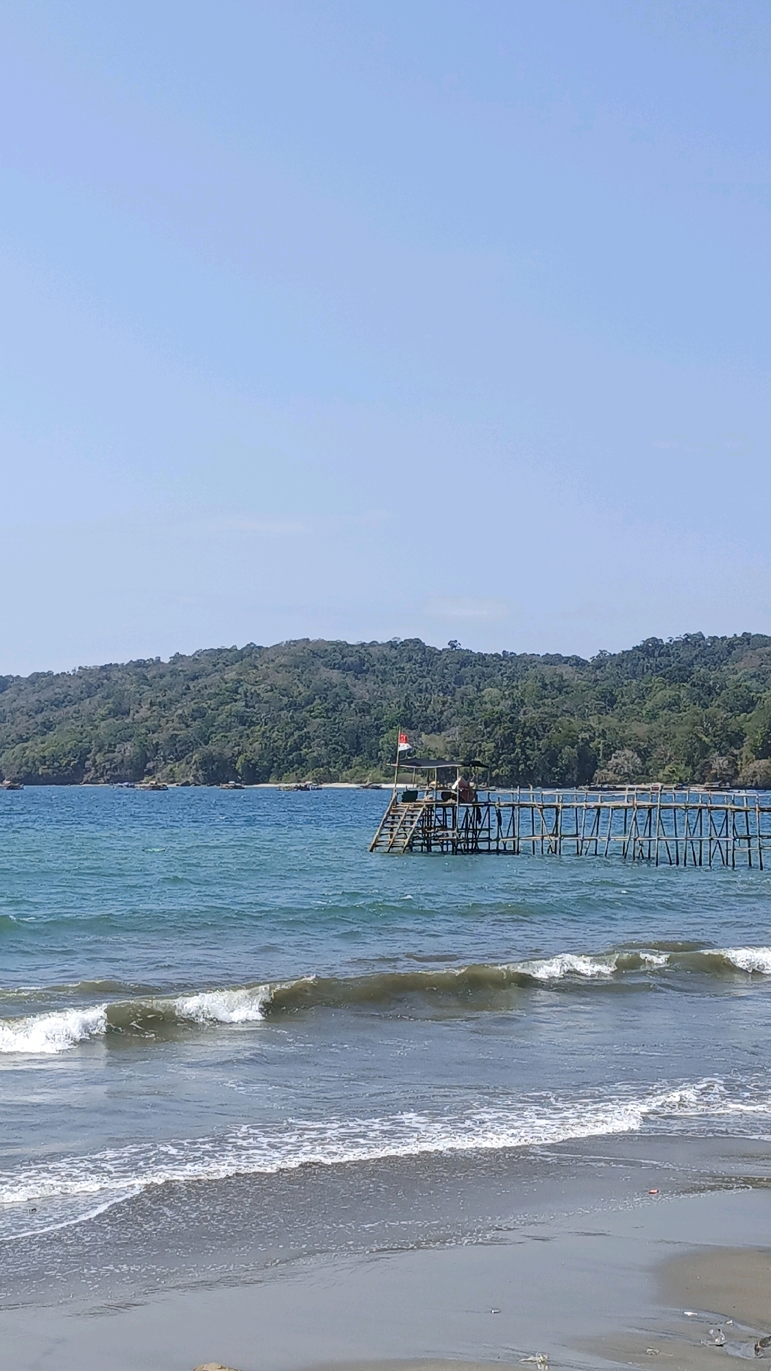 This is a full shot of a beach in Desa Pagandaran, Indonesia. The sky is clear and blue, with no clouds. The ocean is a deep blue, with gentle waves lapping at the shore. In the foreground, there is a sandy beach with some small rocks. A wooden pier extends out into the water, with a small hut on it. An Indonesian flag is flying from the hut. There are two people standing on the pier. In the background, there is a lush green hill covered in trees.