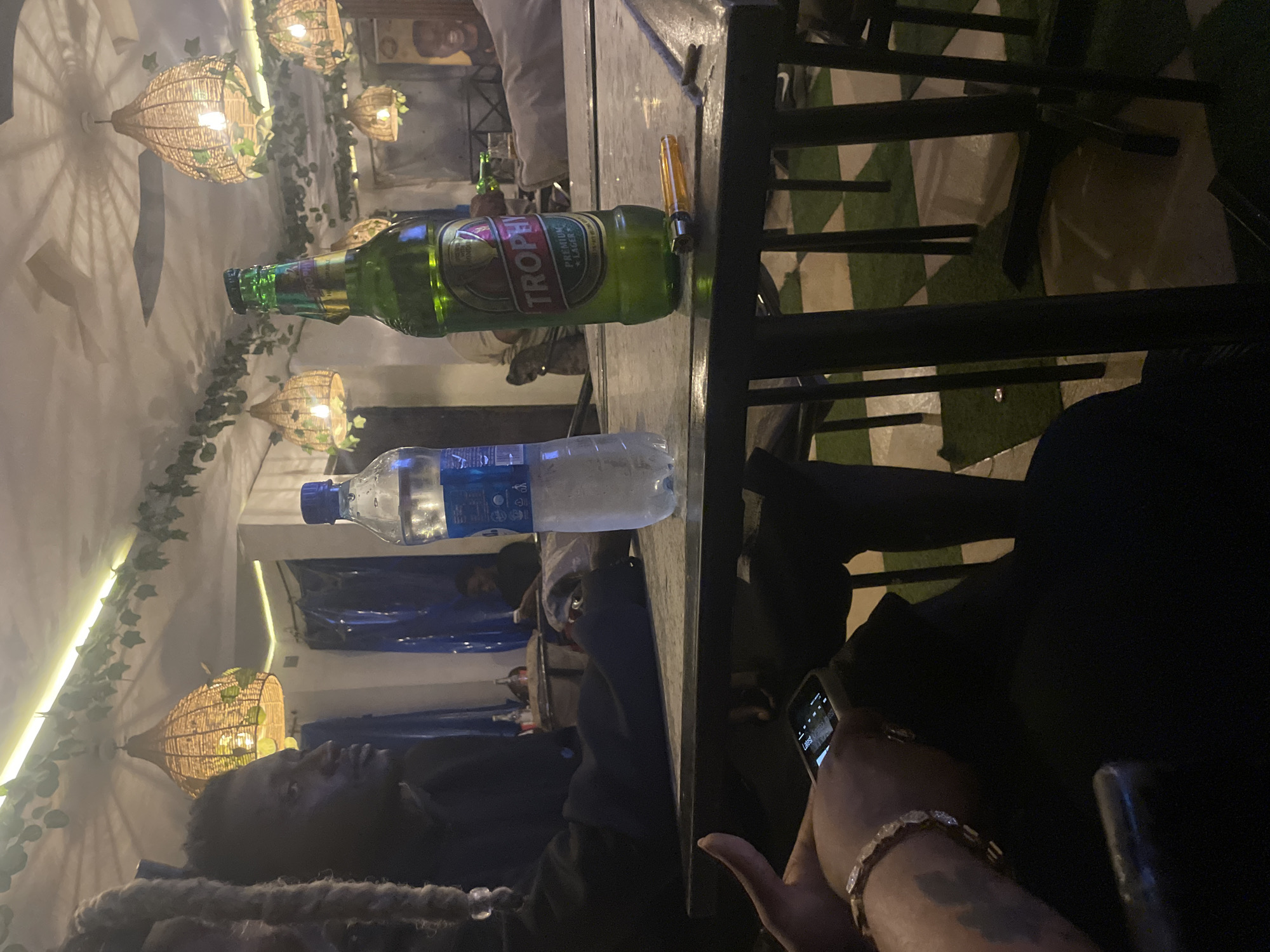 The image captures a scene in what appears to be an outdoor bar or lounge setting in Jos, Nigeria. The camera is positioned at table level, capturing several elements of the environment and people.
Objects on the table include a bottle of Trophy Lager beer and a bottle of water. A metallic bottle opener rests beside the beer.
A person with dark hair and a dark shirt is visible in the foreground, their attention directed towards a phone in their hand. A gold bracelet adorns their wrist.
In the background, another individual is visible.
The setting is decorated with hanging, wicker-like light fixtures intertwined with greenery. The floor appears to be covered with green and white checkered tiling. The lighting is dim, suggesting it is evening or a dimly lit indoor setting.