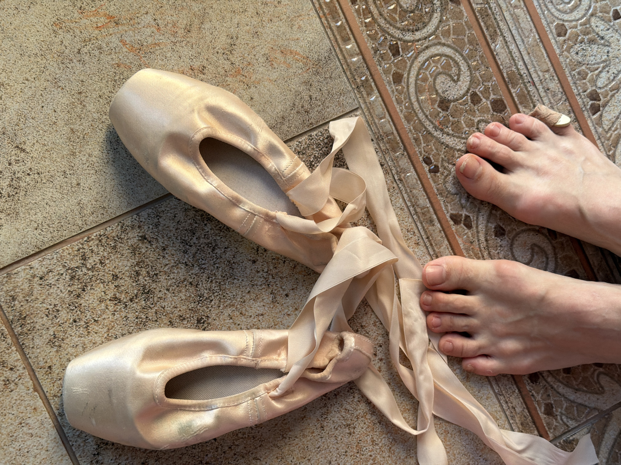 This is an eye-level, medium shot of a pair of ballet pointe shoes with ribbons lying on a tiled floor. A human foot is visible in the lower portion of the frame, with toes pointing towards the shoes. The pointe shoes are made of pale pink satin and are positioned with the soles facing up. The ribbons are untied and spread across the floor. The tiled floor has a beige and brown speckled pattern with a decorative border. The scene is indoors, possibly in a residential setting. The lighting suggests it is daytime, with sunlight casting shadows. There is no discernible indication of the city of Ufa, Russia, or any specific urban environment.