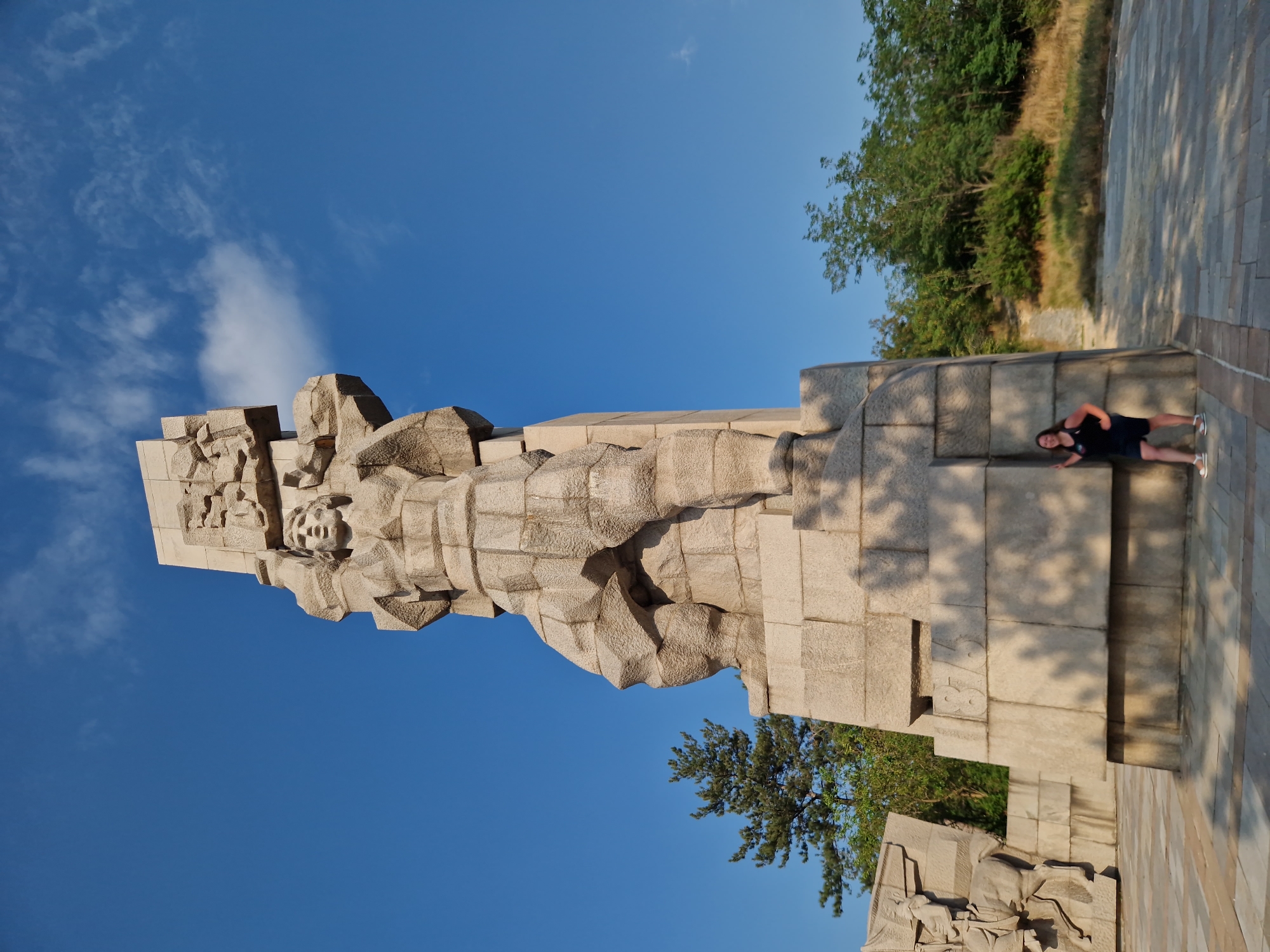 A low-angle, full-shot photograph captures a monumental stone sculpture against a clear blue sky with scattered clouds. The sculpture is a towering, abstract representation of a warrior figure, carved from rough-hewn blocks of stone, with intricate details on the face and body. The figure is depicted in a dynamic pose, holding what appears to be a shield or a large object above its head. The sculpture is situated on a tiered stone base, and in the foreground, a young woman in a black tank top and dark shorts stands leaning against the base, posing for the camera. To the left of the main sculpture, a fragment of another stone relief depicting a horse and rider is visible. The surrounding environment includes a paved area with stone tiles, dense green trees and bushes on a gentle slope in the background, and dry grass. The sculpture is identified as the Monument of the Mother and the Red Army, part of the Panagyurishte Memorial Complex, located in the city of Panagyurishte, Bulgaria, commemorating the 1944 liberation of Bulgaria.