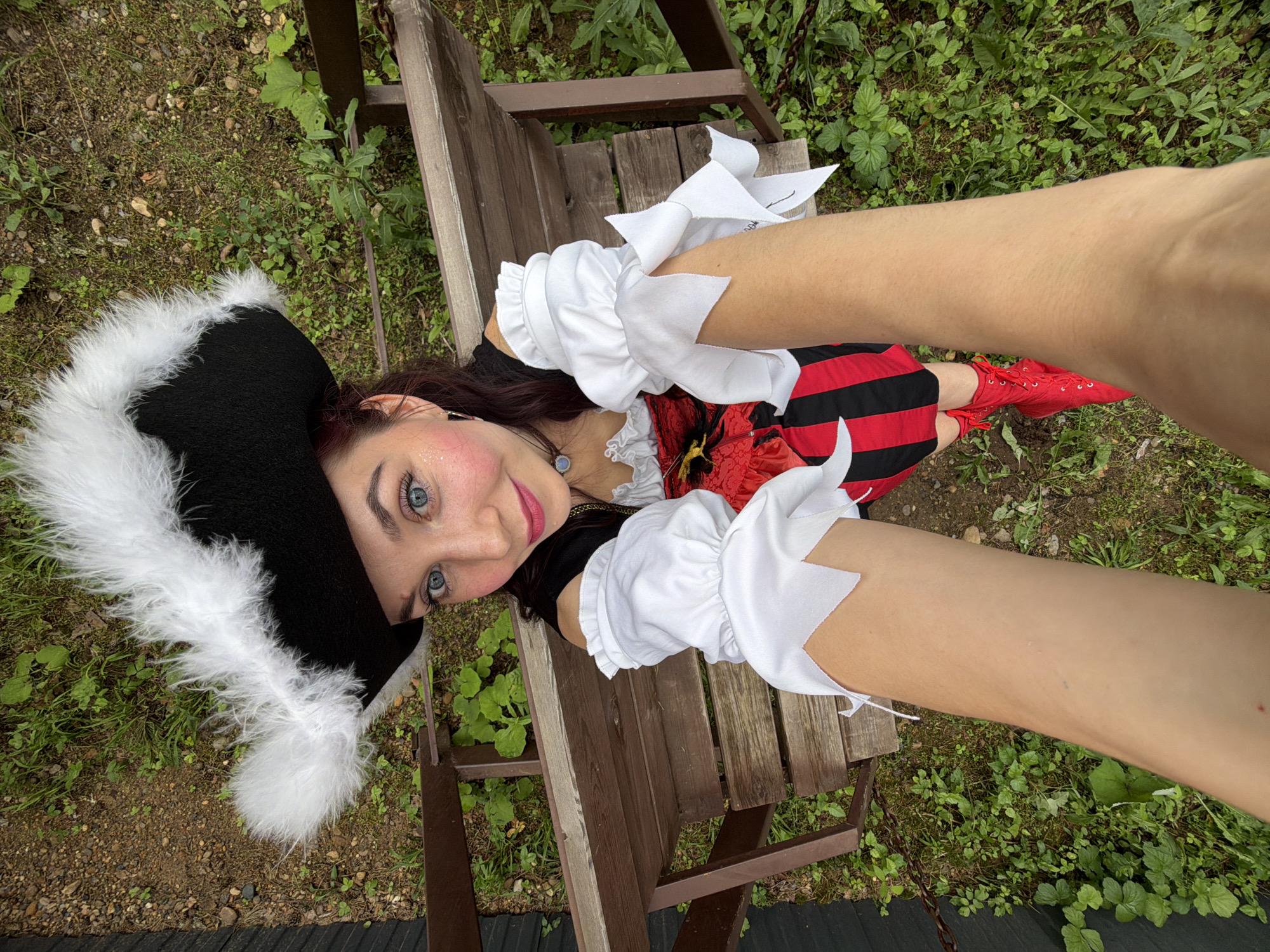 A person is lying on a wooden bench outdoors, wearing a pirate costume. The costume includes a black tricorn hat with white trim and pom-poms, a red and black striped dress, white ruffled cuffs, and red lace-up boots. The person has striking blue eyes and is smiling. The background consists of green foliage and brown wooden planks, suggesting an outdoor setting possibly in a park or garden. The lighting suggests it is daytime. There is no visible text in the image.