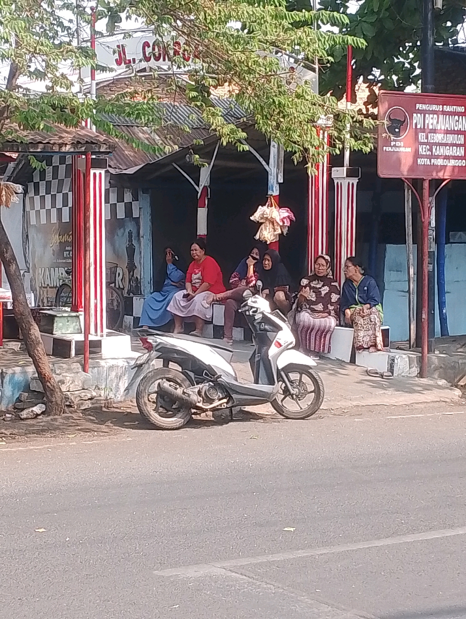 The image shows a street scene in Probolinggo, Indonesia, with several people sitting on a raised platform in front of a building. A white scooter is parked on the side of the road in the foreground.
In the background, a sign is visible that reads "JL. CO..." and the logo of PDI Perjuangan, a political party in Indonesia. Below the party logo, the sign details the location as "KEL. KEBONSARI KULON, KEC. KANIGARAN, KOTA PROBOLINGGO". This indicates the scene is in Kebonsari Kulon Village, Kanigaran District, Probolinggo City.
There are five women seated on the platform. The woman on the far left is wearing a blue patterned dress and a headscarf. Next to her, a woman in a red t-shirt and a light-colored skirt is looking to her right. To her right, a woman in a black hijab and dress is looking to her left. Further to the right, a woman in a patterned brown and white top and a striped skirt is looking forward. The woman on the far right is wearing a blue jacket over a patterned skirt and is looking to her left.
The building behind the women has a corrugated metal roof and red and white striped pillars. There is a checkered pattern on the wall to the left, with the word "KAMPUNG" visible.
The scooter in the foreground is white and appears to be a common model in Southeast Asia. The road in front of the platform is paved and shows signs of wear. The overall atmosphere suggests a casual gathering on a sunny day.