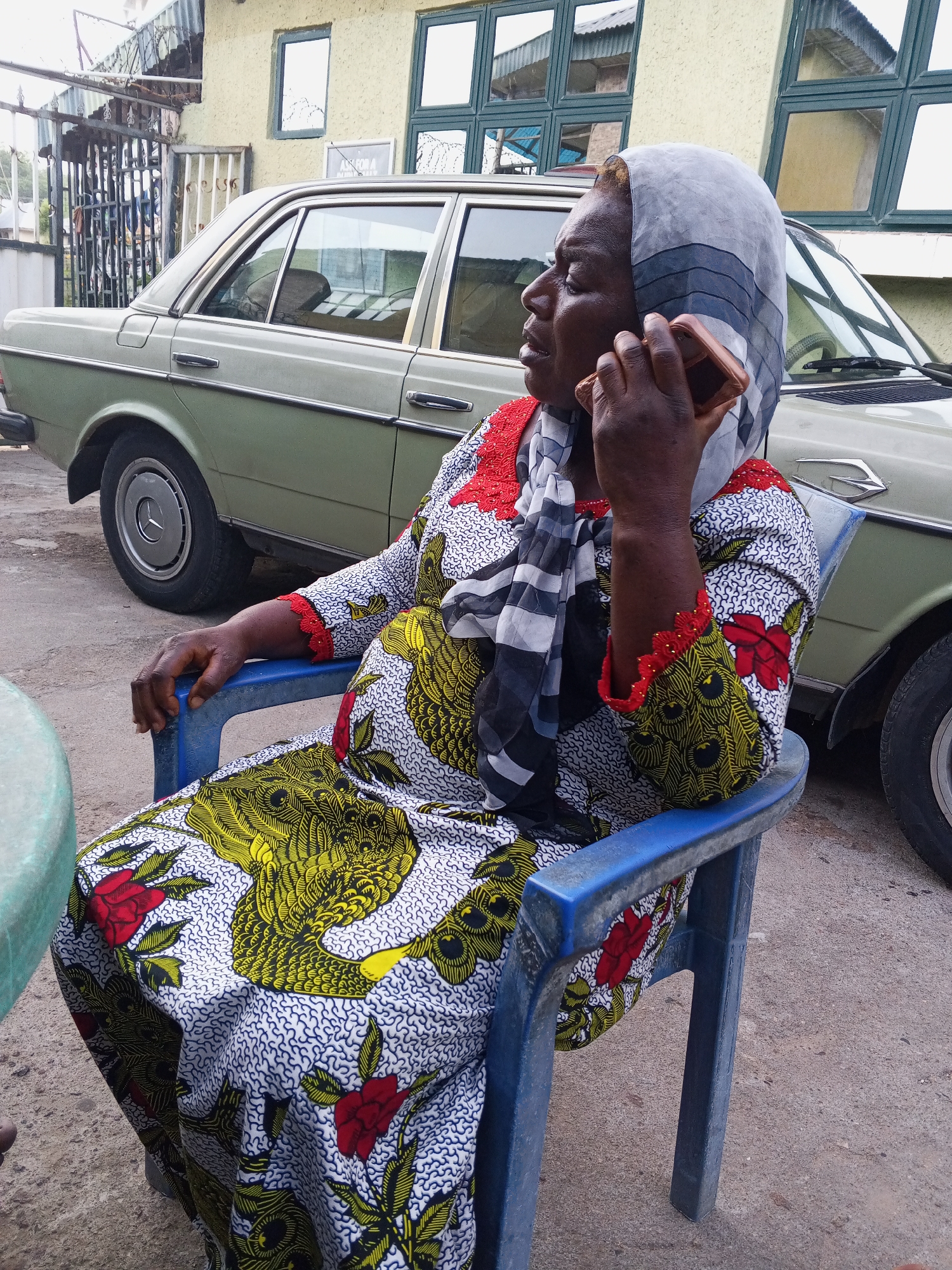 An adult woman is captured in a mid-shot, seated in a blue plastic chair outdoors, engaged in a phone conversation. She holds a rose gold-colored smartphone to her right ear, with her left hand resting on the chair's armrest. Her expression is focused, with eyes slightly closed, suggesting concentration on the call.
She wears a vibrant, patterned dress, commonly known as Ankara fabric, featuring red flowers and prominent yellow/green abstract designs on a white background. Her head is covered with a grey and black striped headscarf.
The setting appears to be a residential or public outdoor space in Zango-ndaji, Nigeria, under natural daylight conditions, possibly overcast. In the background, a light green/beige vintage-style sedan car, identifiable by a visible Mercedes-Benz logo on its wheel hub, is parked. Behind the car, a building with textured, light yellow walls and several dark-framed windows is visible. A metal gate structure can be seen on the far left. Part of a green round table is visible in the foreground on the left. The ground beneath is concrete or asphalt. A faintly visible and largely illegible sign is on the wall behind the car. The overall scene depicts a mundane, everyday moment.