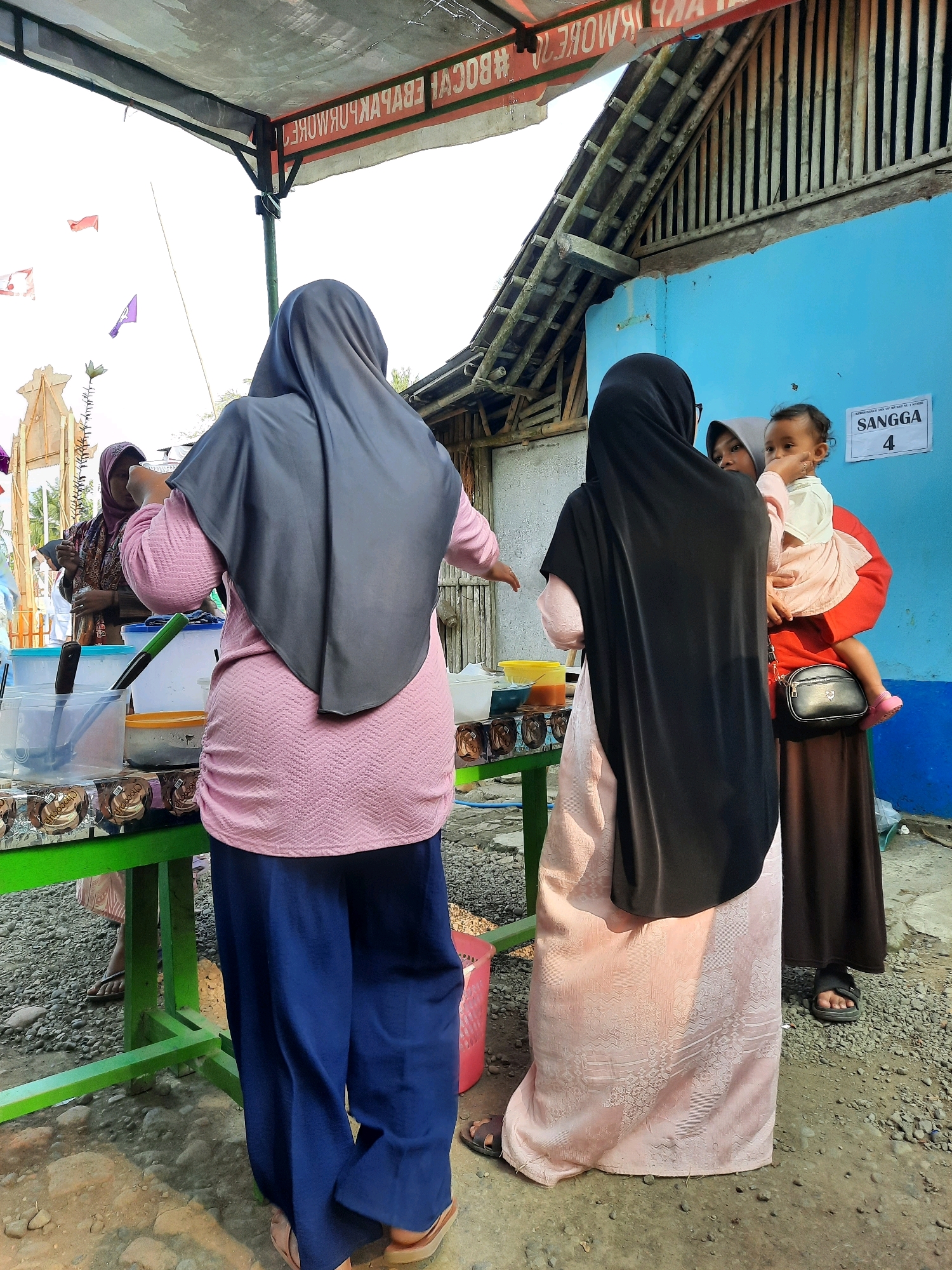 In the city of Suren, Indonesia, at a street-side stall, a scene unfolds under a makeshift awning. A woman, viewed from behind, wears a dark grey hijab and a textured pink long-sleeved top paired with loose, dark blue pants. She stands at a green table laden with various containers and what appear to be individually wrapped food items, possibly snacks or sweets. To her right, another woman, also in a dark hijab, holds a young child. The child, wearing a white top and a pink skirt, looks towards the viewer, its hand near its mouth. The woman holding the child is dressed in a light pink, patterned dress and a brown skirt. Behind them, a bright blue wall serves as a backdrop, adorned with a white sign that reads "SANGGA" followed by the number "4". The ground is a mix of gravel and dirt, with a few scattered rocks. The overall setting suggests a casual, outdoor market or community event.