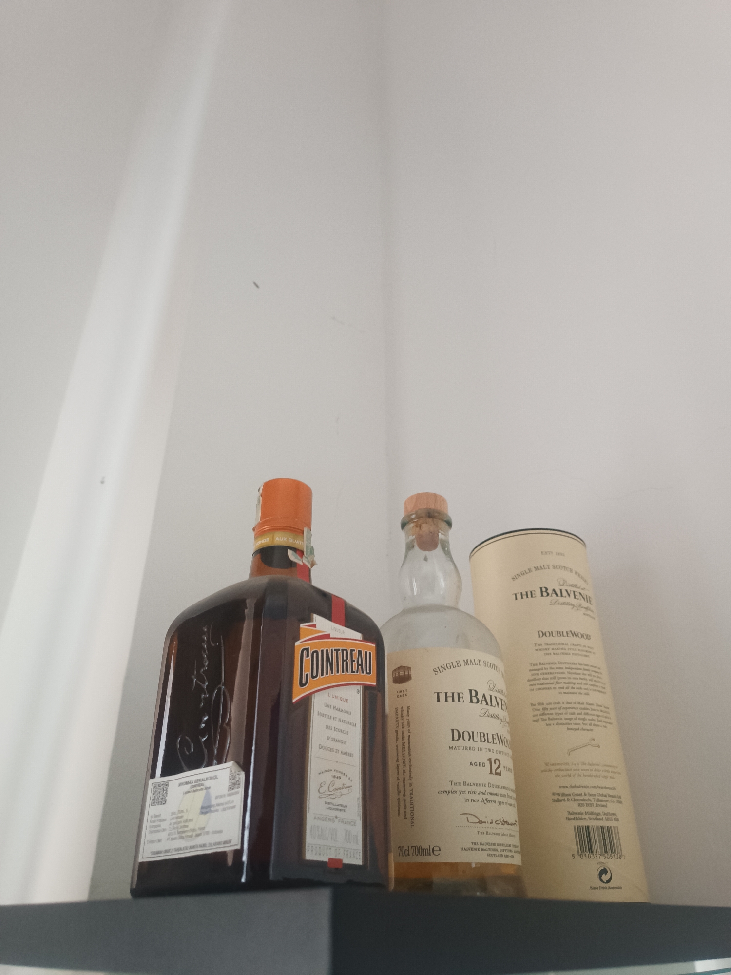 The image shows three bottles on a shelf. The bottle on the left is a brown glass bottle with an orange cap and a label that says "COINTREAU". The bottle in the middle is a clear glass bottle with a cork stopper and a label that says "SINGLE MALT SCOTCH WHISKY". The bottle on the right is a tall, light-colored cylinder with a label that says "THE BALVENIE DOUBLEWOOD AGED 12 YEARS". The background is a plain white wall. The image is taken from a low angle, looking up at the bottles. The lighting is bright and even. There are no people or animals in the image.