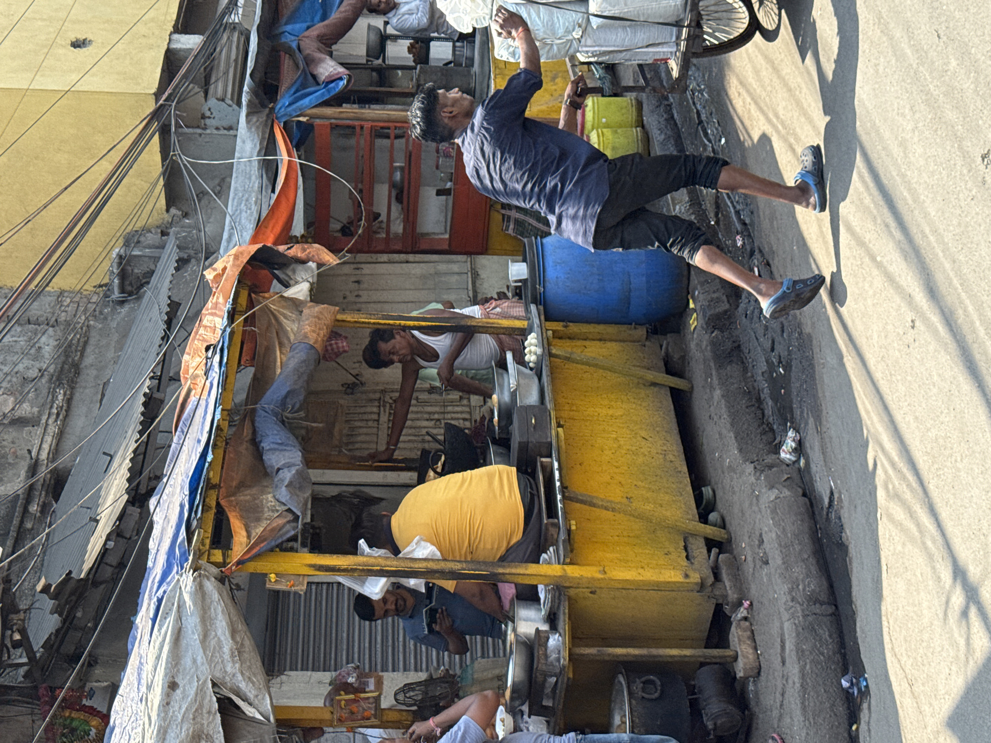 The image shows a street scene in Kolkata, India. A man in a blue shirt and black pants is reaching up toward the top of a food cart, which is constructed of yellow wood and covered in blue and white tarps. The cart appears to be a vendor's stall, with several people inside preparing food. One man is wearing a white tank top and working with large metal bowls. Another man is wearing a yellow shirt. A blue barrel is visible next to the cart. Other people are resting or working within the cart's confines. Overhead, there are numerous power lines. On the street, there is refuse and shadows cast by the sun.