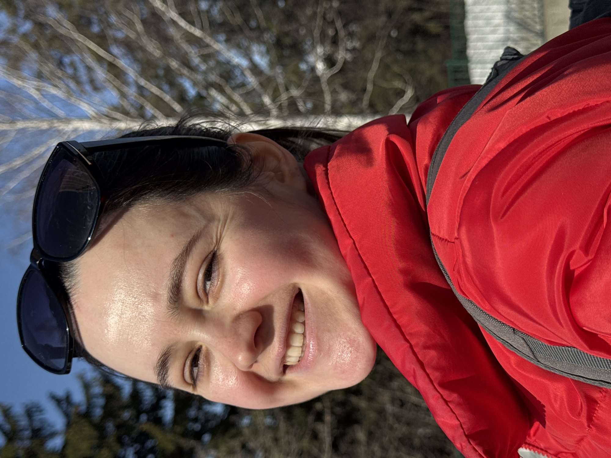 A person with fair skin and dark hair smiles at the camera. Sunglasses are pushed up on their head. They wear a red jacket with gray trim. The background shows bare trees and a blue sky. There is not enough information to determine if the location is Izhevsk, Russia.
