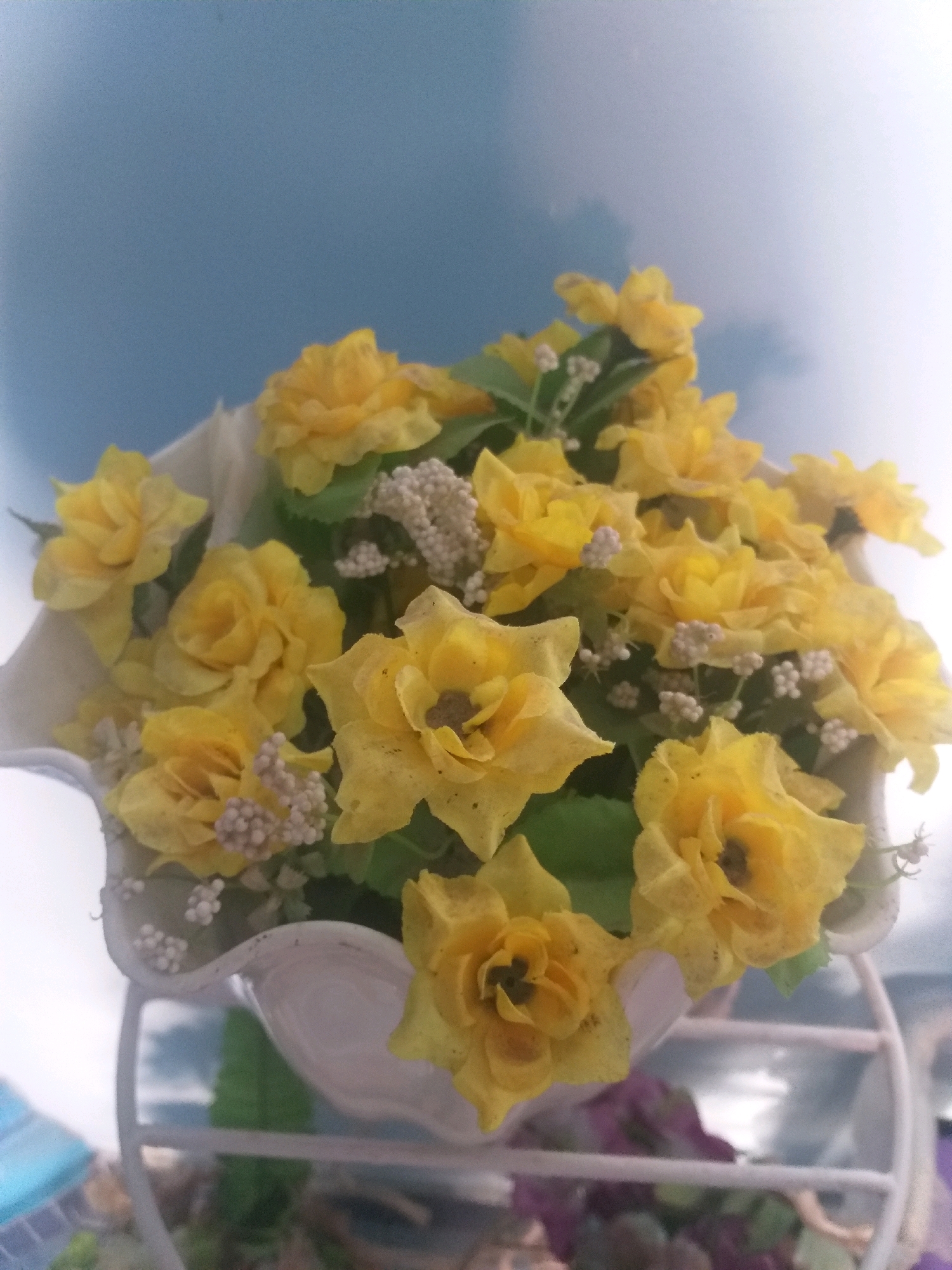 A bouquet of artificial yellow roses is displayed in a white ceramic pot, which is set on a white metal stand. The background is a blurry blue sky with some lighter clouds. The lighting suggests it is daytime. The scene is indoors, near a window, with hints of what might be other objects or furniture visible in the lower portion of the image. There are no people or animals present. The image does not provide any specific information about the city of Dumai, Indonesia.