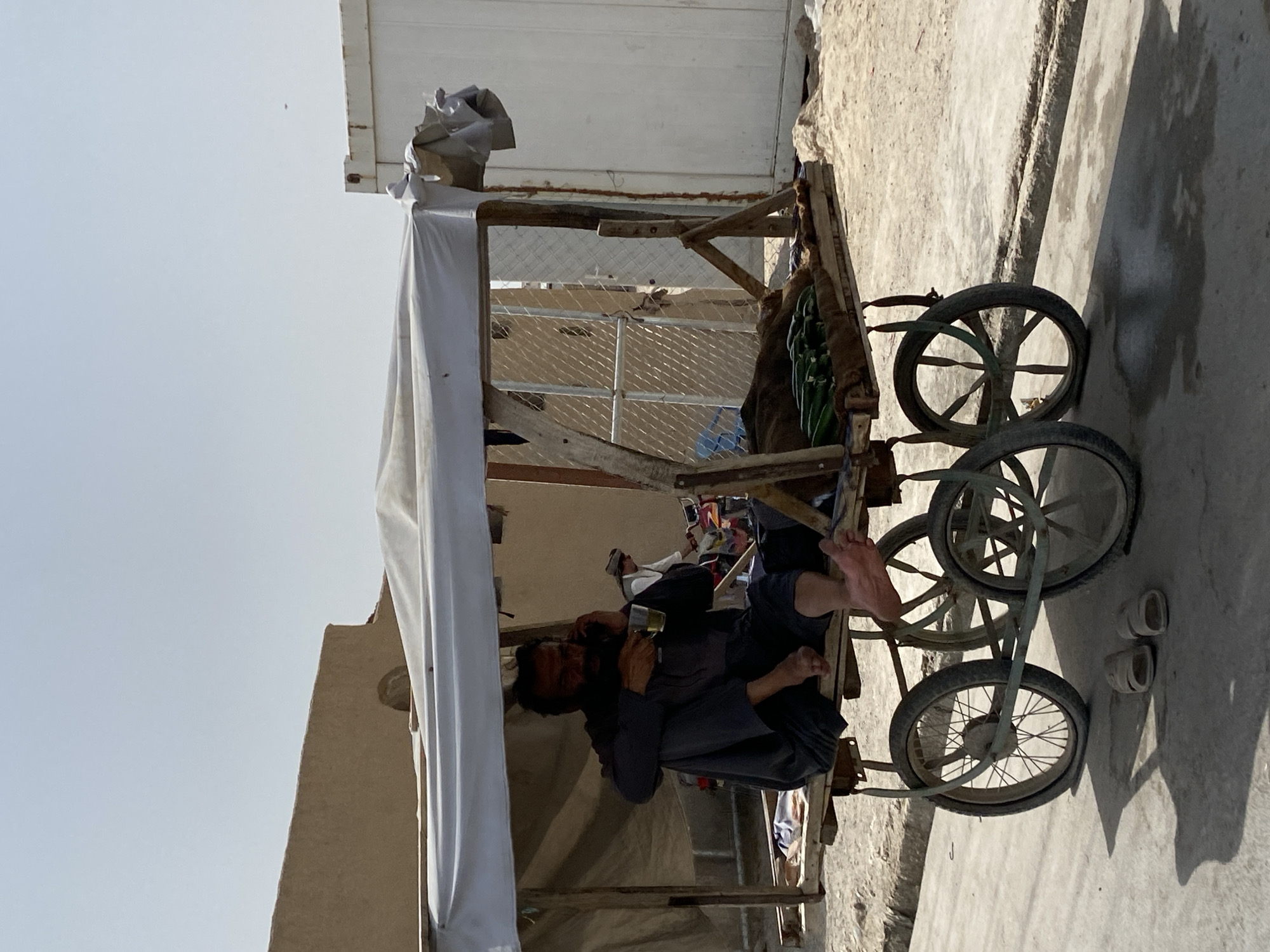 A man is sitting on a wooden cart with a canopy, holding a cup and a phone, in what appears to be the city of Kandahar, Afghanistan. The cart is loaded with cucumbers. The man is wearing a dark blue garment. The cart has two large wheels at the back and two smaller wheels at the front. A pair of sandals is on the ground next to the cart. In the background, there are buildings and shipping containers, suggesting an urban or semi-urban environment. The sky is clear and bright.