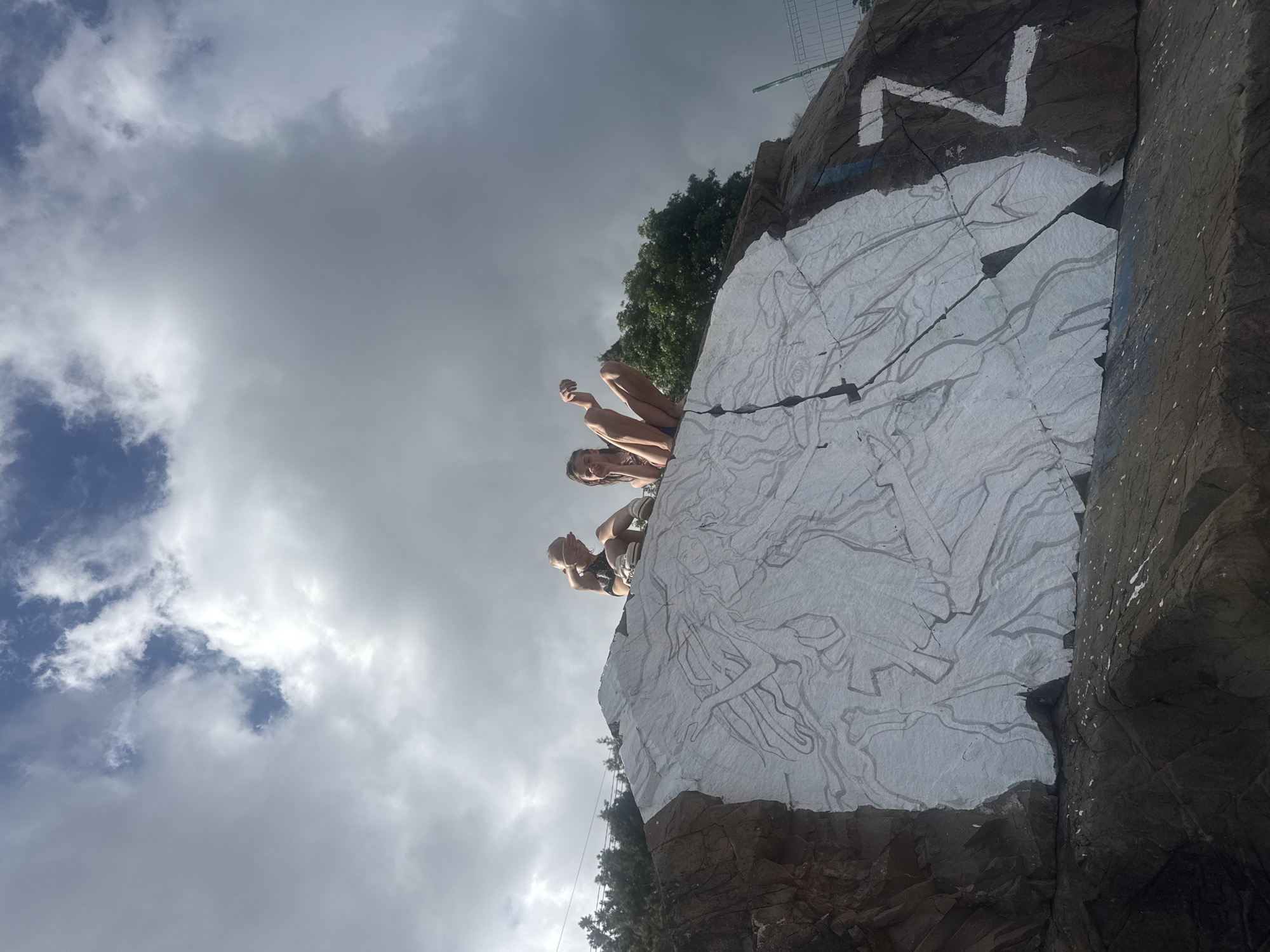 Two young females are sitting on the edge of a rocky outcrop. The rock face has a large white mural painted on it with grey line drawings of figures. To the right of the mural, a large white "Z" is painted on the rock. In the background, there are green trees and a blue sky with white and grey clouds. The scene appears to be outdoors, possibly in a natural or semi-natural setting within the city of Partenit, Ukraine.