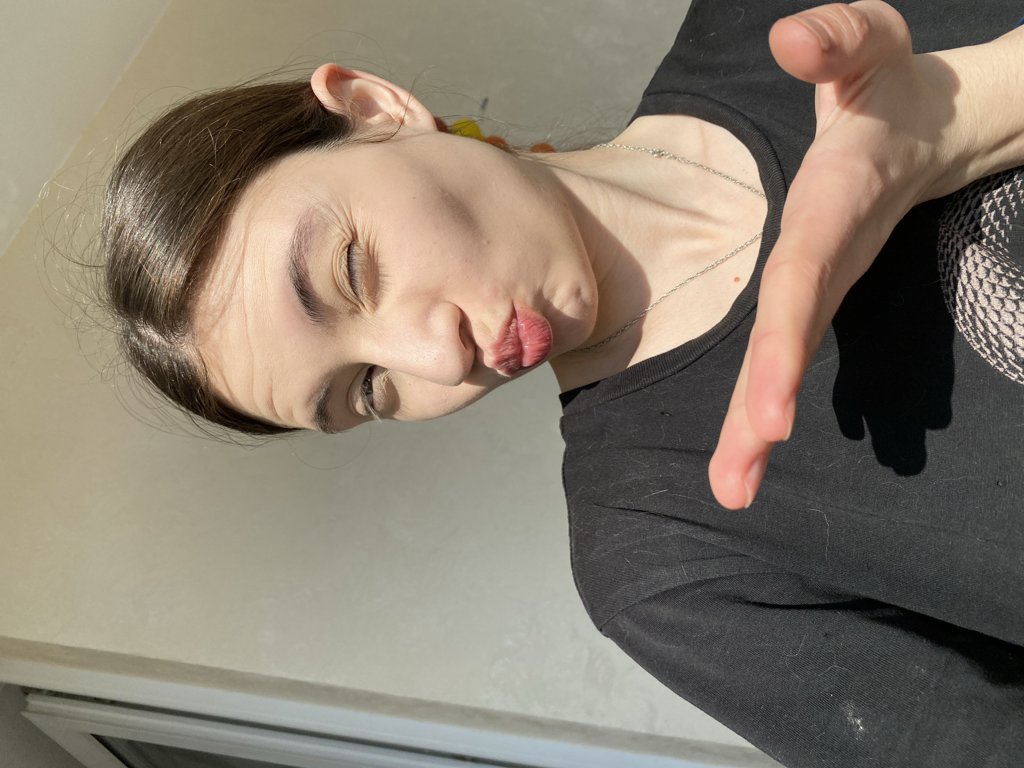 A person with dark brown hair, light skin, and a silver chain is looking downward, with eyes partially closed and lips pursed. The person is wearing a black shirt and their right hand is outstretched towards the camera. The background is a light beige wall with a partially visible window frame. There is no specific information to identify the image's location in Votkinsk, Russia.