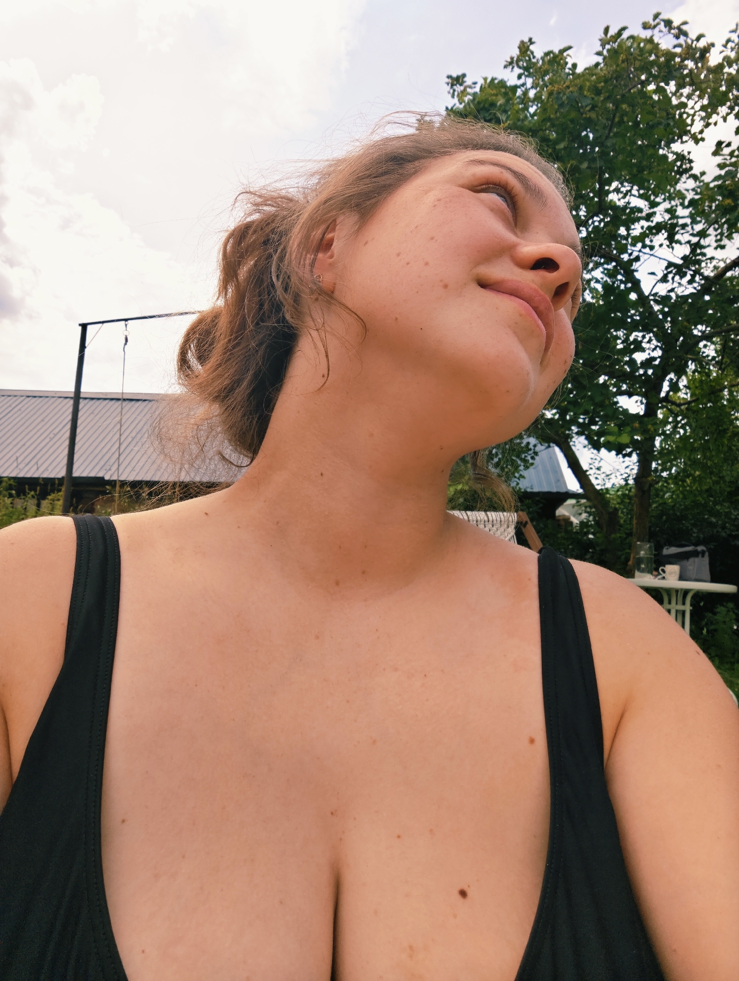 A medium shot taken from a low angle shows a young woman with light skin and freckles looking up and to the right, with a slight smile. Her light brown hair is pulled back in a messy bun, and she is wearing a black tank top, revealing her chest and shoulders. Her skin appears sun-kissed. The background is slightly blurred, showing a building with a corrugated metal roof and a tall tree with green leaves. The sky is cloudy and bright. The scene appears to be outdoors in a rural or suburban setting, possibly in Noviy, Russia, given the context provided.
