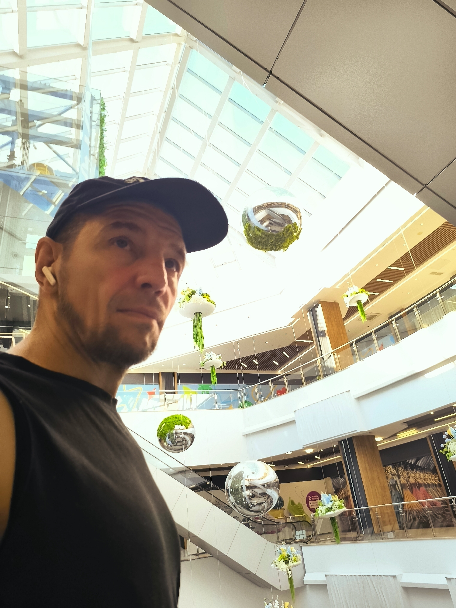 A medium shot captures a man wearing a black baseball cap and a black sleeveless shirt, looking off to the right. He has a light beard and is wearing white wireless earbuds. The man is positioned in the foreground of a modern indoor atrium, likely a shopping mall, characterized by its multi-level structure and glass ceiling. Suspended from the ceiling are several decorative spheres, some containing green moss or foliage, and floral arrangements. The background reveals escalators, glass railings, and large artistic murals or advertisements on the walls of the upper levels. The scene is brightly lit, suggesting daylight filtering through the glass roof. The location is identified as Qarağandı city administration, Kazakhstan.