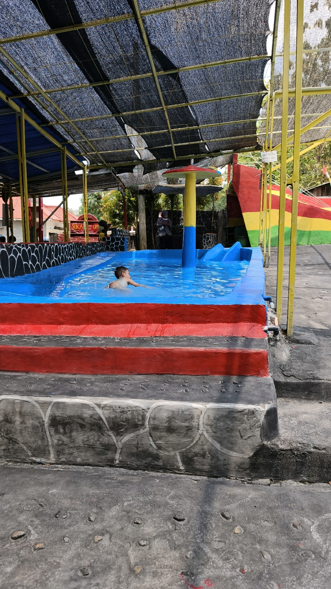 A young person swims in a blue swimming pool. The pool is surrounded by red and blue painted concrete steps. A yellow and blue mushroom-shaped structure with a red cap stands at the edge of the pool, next to a red and yellow slide. A black mesh shade covers part of the area, supported by yellow metal poles. In the background, trees and buildings are visible. This scene is likely located in Air Paoh, Indonesia, given the context.