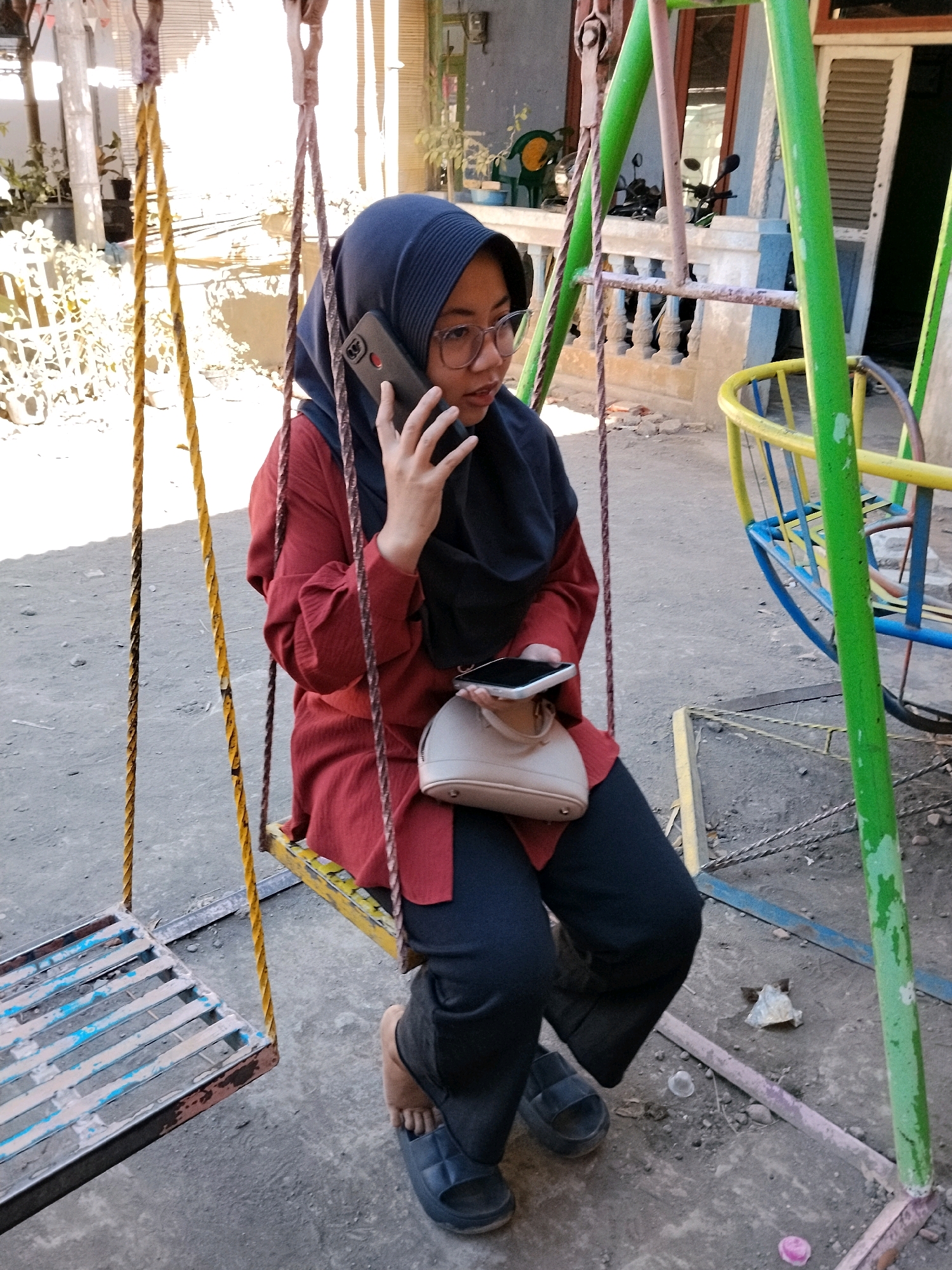 A young woman wearing a navy blue hijab, glasses, and a red long-sleeved shirt is sitting on a swing, holding a smartphone to her ear. She is also holding another smartphone in her left hand. A beige handbag rests on her lap. She is wearing dark pants and dark sandals. The swing set has green and yellow metal supports and yellow and brown ropes. The background shows a courtyard with concrete ground, a building with windows and doors, and some plants. The scene appears to be outdoors in a residential area.