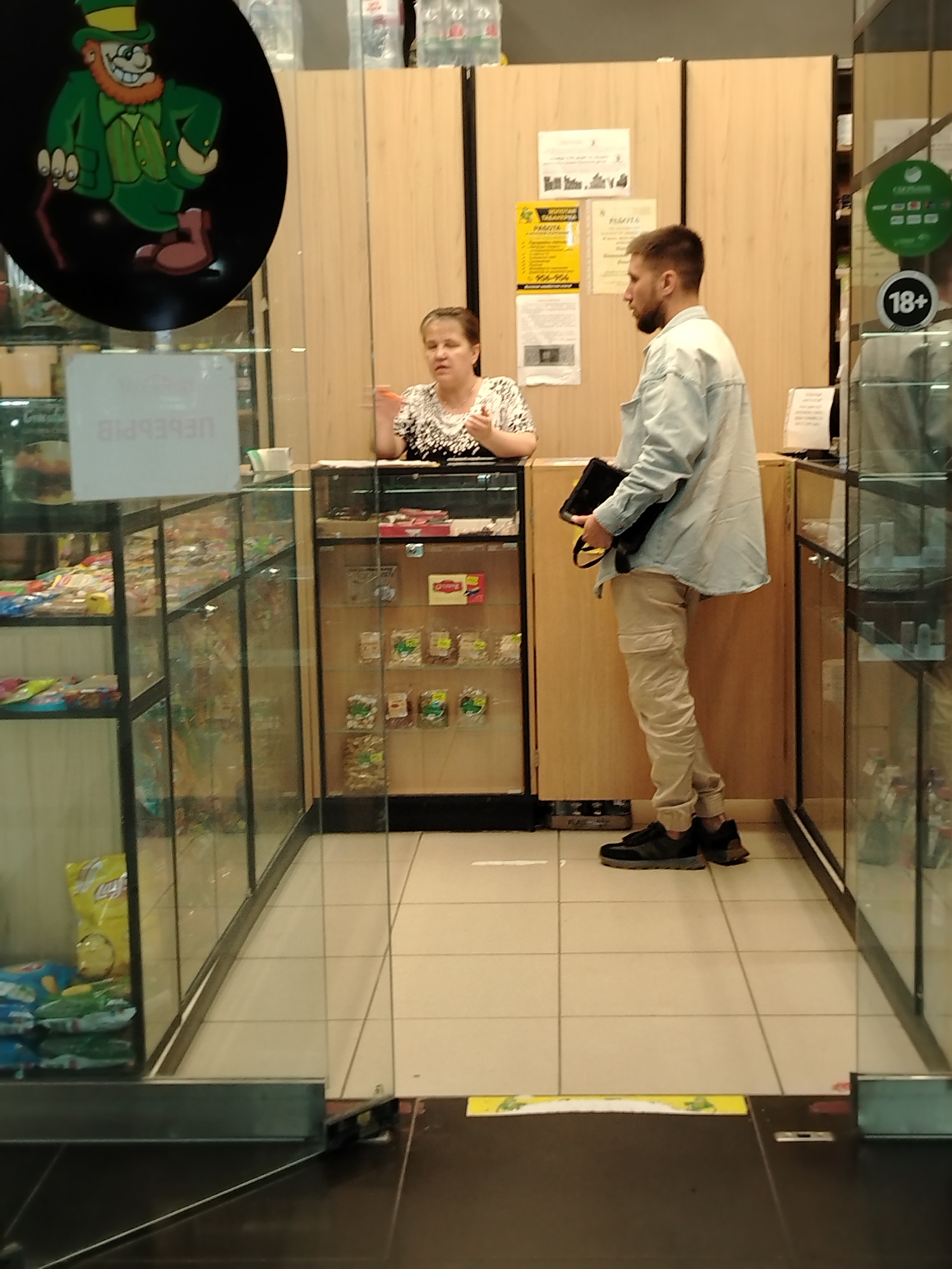 This image captures a customer interaction at a small retail kiosk or shop, likely in Izhevsk, Russia.
**Subjects:**
A woman, appearing to be a shop attendant or cashier, stands behind a wooden counter. She has dark hair, wears a dark top with a white pattern, and is gesturing with her right hand while holding a pen-like object, looking towards the customer.
A man, likely a customer, stands facing the attendant. He has short hair and a beard, and is wearing a light blue or white oversized button-up shirt, light beige cargo pants, and dark sneakers. He holds a black bag or tablet in his left hand.
**Scene and Setting:**
The setting is an indoor retail space, characterized by glass display cases filled with various packaged snacks, candies, and other small items, flanking the customer area. A prominent yellow "Lay's" chip bag is visible in the left display case. The counter behind the attendant is wooden, and the wall behind her is also wooden, featuring several papers and signs. The floor is made of light-colored square tiles. The overall impression is a compact, functional retail point.
**Activity:**
The image depicts a routine customer service interaction, with the customer and attendant engaging in what appears to be a transaction or inquiry.
**Notable Details:**
Artificial lighting illuminates the scene, typical of an indoor commercial environment. The mood seems neutral, focused on the exchange. A large, circular black sign on the left features a cartoon leprechaun character in a green suit, top hat, and holding a cane. Another circular sign on the right displays "18+", indicating age-restricted products are sold.
**Visible Text:**
* On the white rectangular sign in the foreground (possibly reflected on glass): "ПЕРЕРЫВ" (Pereryv), which means "Break" in Russian.
* On a yellow sign affixed to the wooden wall behind the man: "РАБОТА" (Rabota), meaning "Work" or "Job," suggesting an employment advertisement. A partial phone number "956-956" is also visible.
* The "18+" sign, indicating an age restriction.
* The brand name "Lay's" on a chip bag.