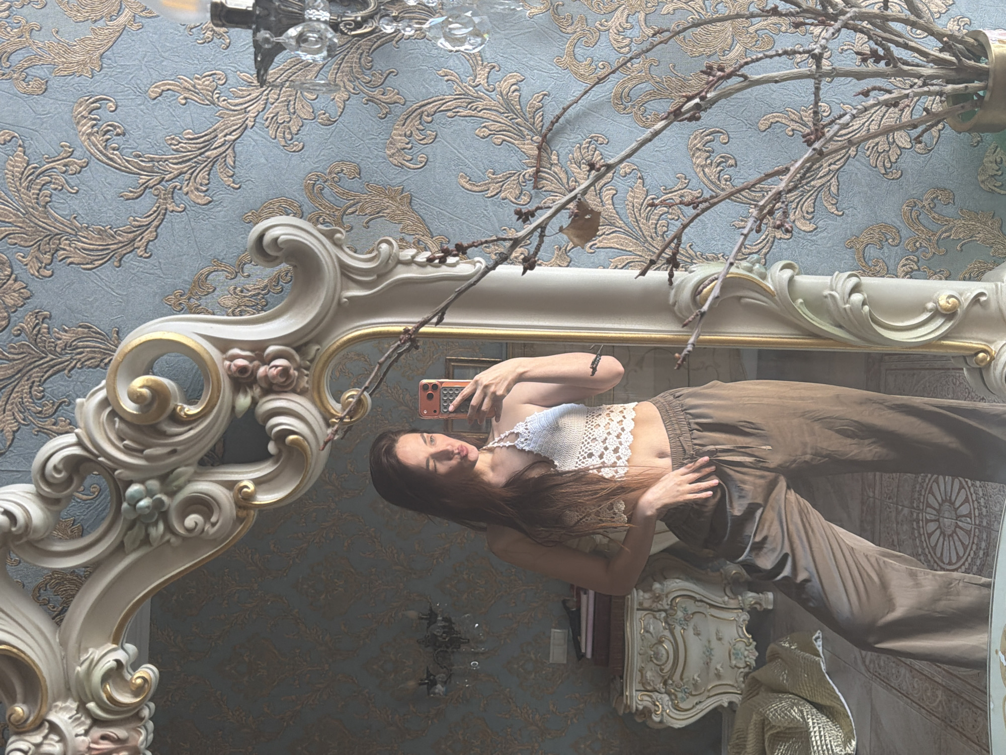 This is a vertically oriented, full-body mirror selfie taken in a room with ornate, vintage decor. The subject is a young woman with long, dark hair, wearing a white crocheted crop top and loose, brown pants. She is holding a smartphone with an orange case in her right hand and is posing by puckering her lips. Her left hand rests on her hip.
The background features a light blue wallpaper with a raised, gold damask pattern. A large, ornate mirror with a cream-colored, carved frame dominates the foreground. The mirror's frame has gold accents and decorative floral elements. Branches of a dried plant extend into the frame from the top, casting shadows.
The reflection in the mirror shows a woman who appears to be in a bedroom or a sitting room. A vintage-style dresser or cabinet is visible to the right of the woman, adorned with similar ornate carvings. A metallic gold blanket or textile is draped over it. There are also some books stacked on a shelf behind the dresser. The lighting suggests it is daytime, possibly late afternoon, with soft light filtering into the room. The overall mood is relaxed and casual. No visible text is present in the image. The location context is Ufa, Russia.