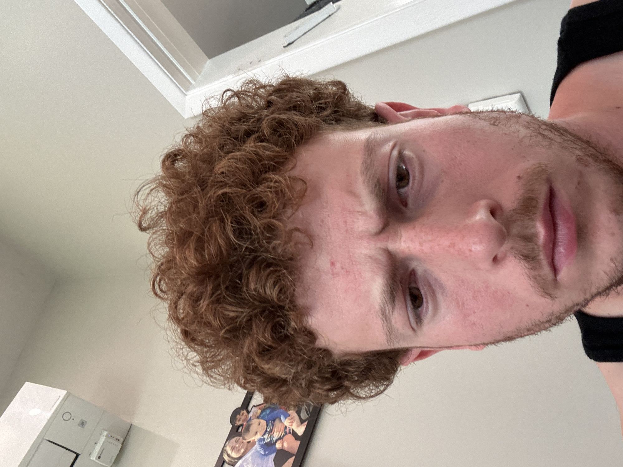 A close-up, low-angle photograph depicts a light-skinned individual with curly, reddish-brown hair. The person's face is visible, showing brown eyes, a mustache, and a light beard. The individual appears to be wearing a black garment at the edge of the frame. The background includes a white ceiling with molding, a framed picture on the wall, and a white electrical panel. No information within the image indicates a location in Pepper Pike, United States of America.