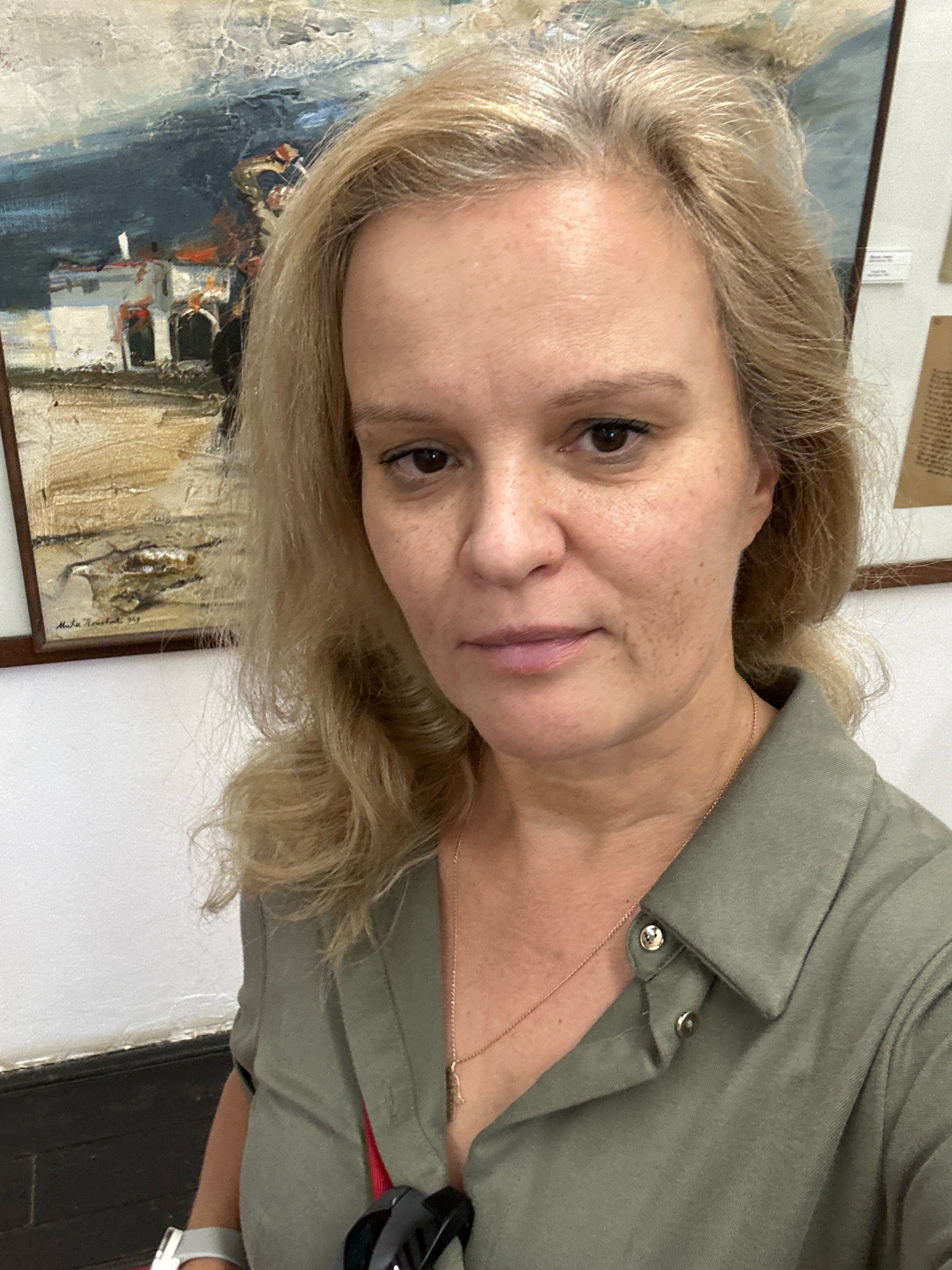 A middle-aged Caucasian woman with blonde hair, fair skin, and brown eyes is taking a selfie. She is wearing a green collared shirt with silver buttons and a thin gold necklace with a pendant. In the background, there is a painting with a landscape depicting buildings and a body of water, and a framed document. The environment appears to be indoors, possibly a museum or gallery. The woman is looking directly at the camera with a neutral expression.