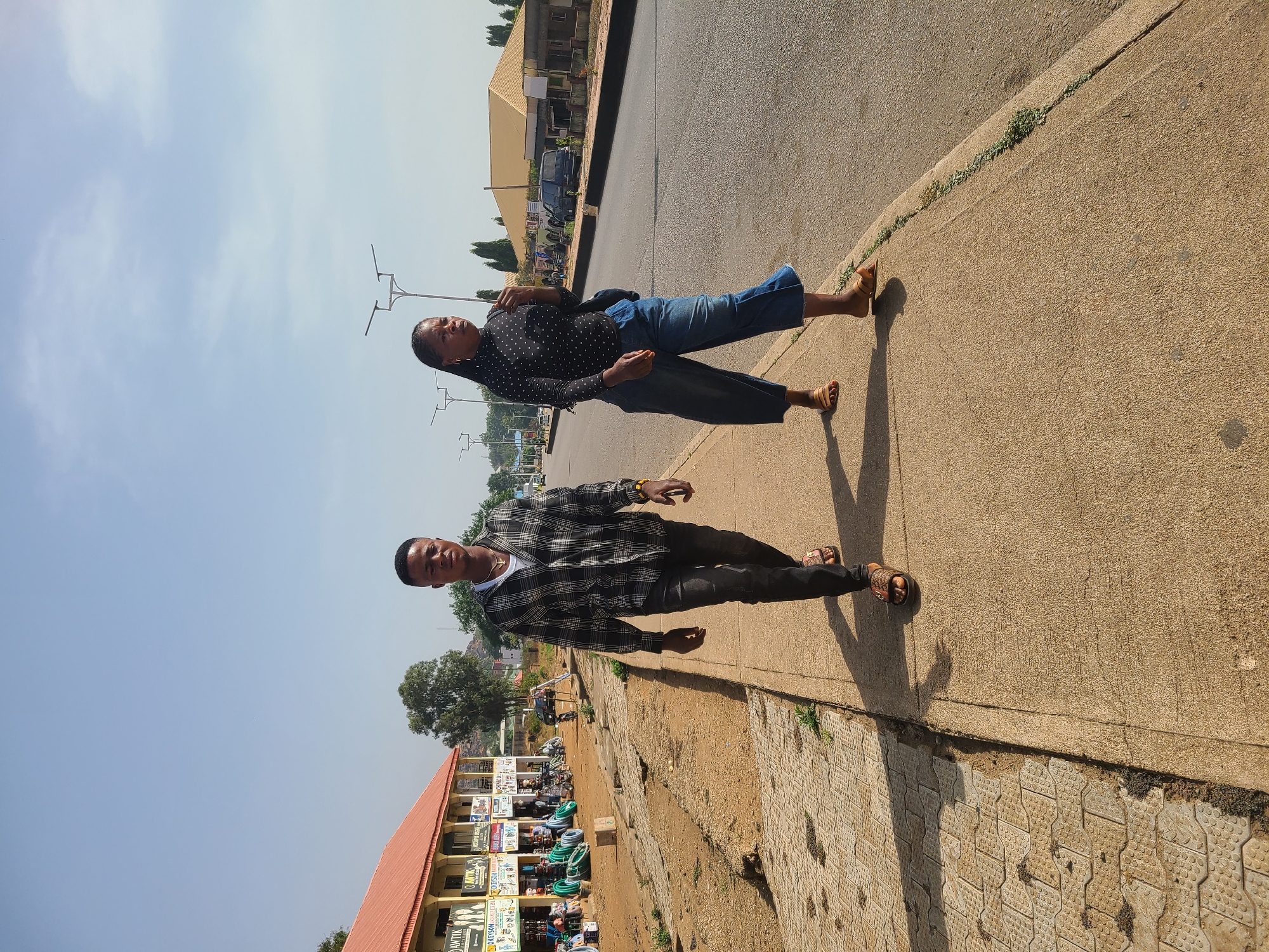 This image, taken in Jos, Nigeria, features two individuals, a man and a woman, standing on a paved sidewalk next to a road. The scene is illuminated by bright daylight under a clear blue sky, with distinct shadows suggesting either mid-morning or late afternoon.
The man, positioned on the left side of the frame, wears a black and white plaid long-sleeved shirt over a white t-shirt, black trousers, and brown sandals. He is looking directly at the camera with a neutral expression. The woman, on the right, is dressed in a black long-sleeved top with white polka dots, blue wide-leg jeans, and brown sandals. She is also looking towards the camera, slightly turned.
The background reveals a bustling street environment with various buildings lining the road, some functioning as shops or stalls. Goods are displayed outside some establishments. Visible text on the signs includes "AKWATA," "BEST CHOICE," "KENT," "DAV," and "FASHION," indicating commercial activity. Trees, sparse vegetation, and utility poles/streetlights are also present along the street. The subjects appear to be posing for the photograph, possibly taking a break from walking along the vibrant street.