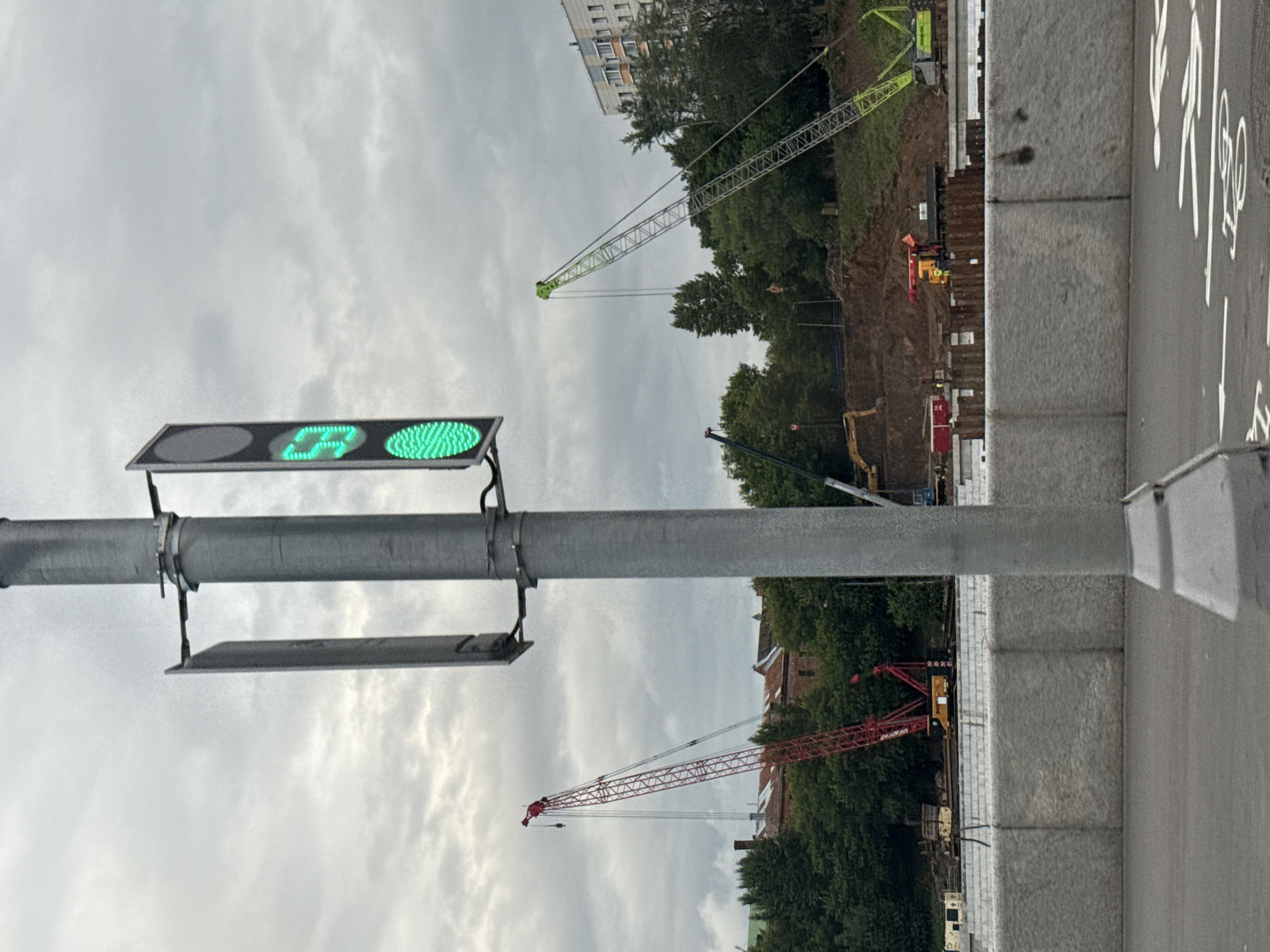 This image captures a street scene in Unknown, Unknown under a cloudy sky. In the foreground, a grey traffic light pole stands prominently, featuring a vertical traffic signal displaying a green "53" and a fully illuminated green circle at the bottom. To the left of the pole, two grey rectangular shields are mounted. Behind the traffic pole, construction is actively underway. Red and green cranes are visible, along with heavy machinery and earth-moving equipment, indicating ongoing infrastructure development. A large concrete barrier runs horizontally across the middle ground, separating the road from the construction site. Trees and buildings are visible in the background, suggesting an urban or suburban setting. White lines, possibly a bike lane, are marked on the road in the lower right portion of the image.