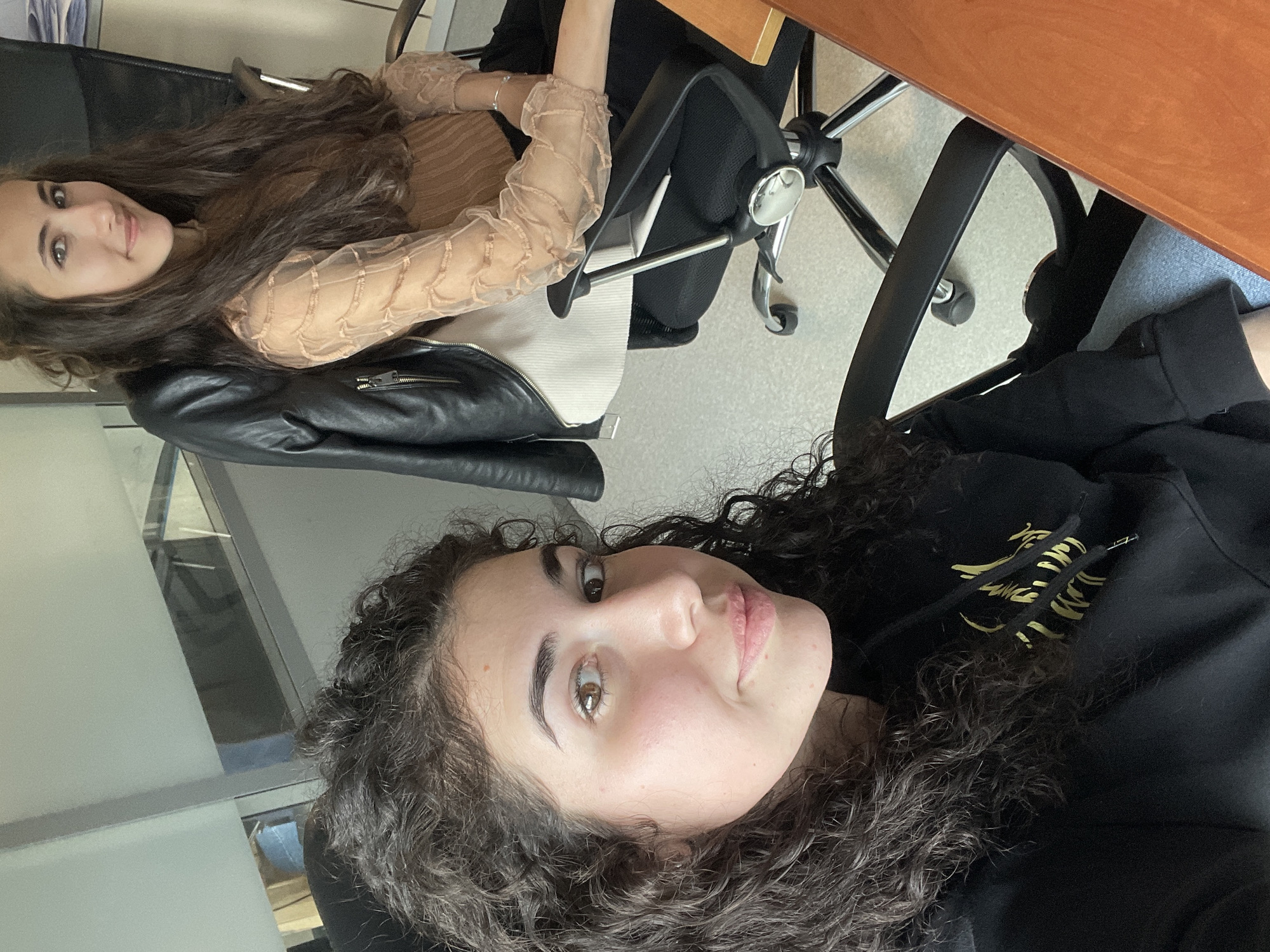 Two women are indoors, likely at an office space. One has long brown hair and is wearing a tan long-sleeved sheer shirt over a white top, with a black leather jacket near her. The other has dark curly hair and is wearing a black hoodie with gold designs. There is a wooden desk visible, as well as office chairs. The location is suspected to be in Sofia, Bulgaria, but there's no definitive visual information to confirm this.