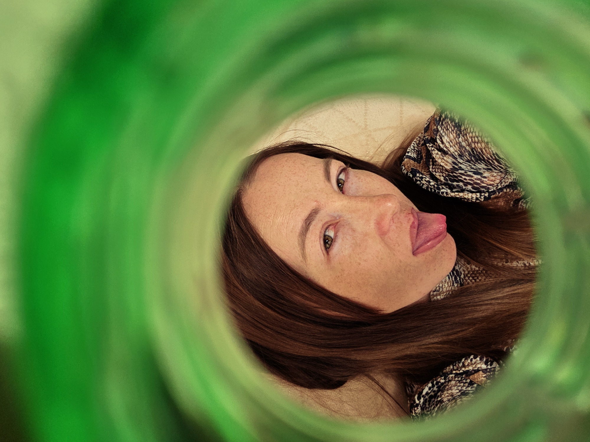 A woman with brown hair and freckles is sticking her tongue out, as seen through a circular green opening. The background is a light beige color. She is wearing a patterned top.