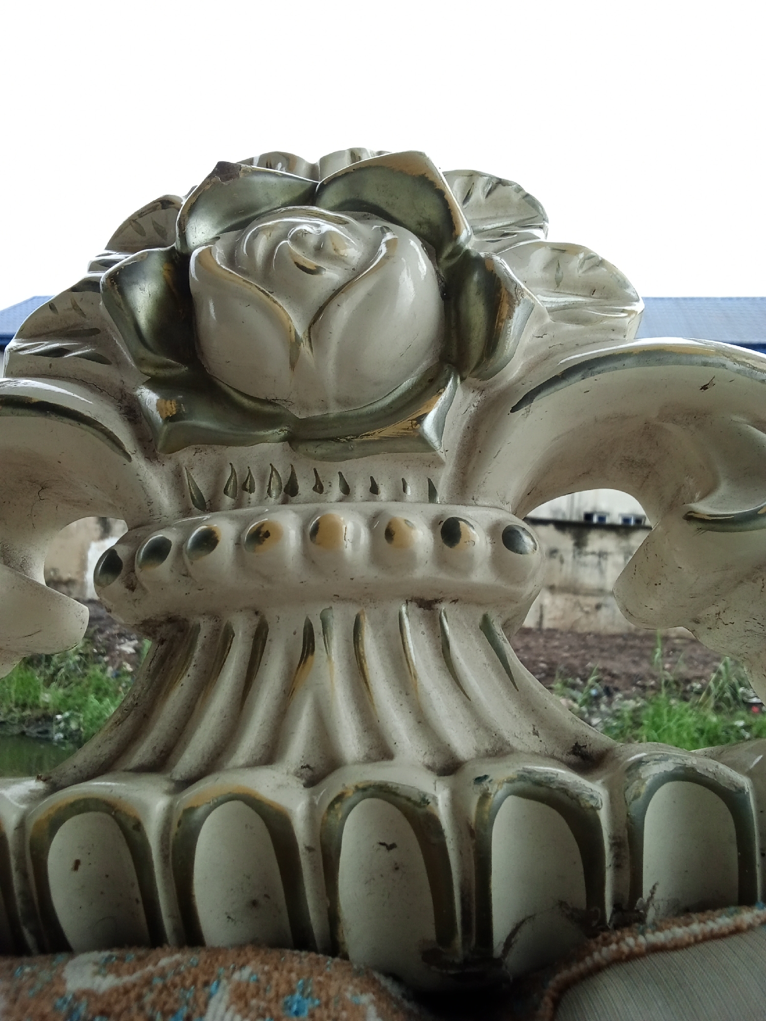 This close-up, vertical photograph captures an ornate decorative object, likely part of a furniture piece or architectural embellishment, situated outdoors in Ado, Nigeria. The primary subject is a sculptural element rendered in off-white or cream, accented with metallic gold or bronze paint that highlights its intricate details.
At the top, a prominent rose motif is surrounded by scrolling leaf-like patterns. Below this, a horizontal band features a series of spherical or domed protrusions, some appearing darker in their centers. Further down, the structure displays vertical fluting and ribbed textures, culminating in a base with distinct arched, recessed sections. The object shows signs of wear, including dirt accumulation, minor chipping on surfaces like the rose petals, and some fading or peeling of the metallic accents, suggesting age and exposure to the elements.
The background is softly blurred, indicating a shallow depth of field. A bright, almost entirely white sky dominates the upper portion, suggesting an overcast day or strong overhead illumination. Visible in the mid-ground are patches of green vegetation, possibly grass, along with areas of dark soil or earth. Faintly discernible in the distance are blurred structures, including a building with a dark roofline and what appears to be a light-colored wall or concrete surface. There are no people, animals, or visible text. The image portrays a static scene focused solely on the decorative object within its outdoor setting.