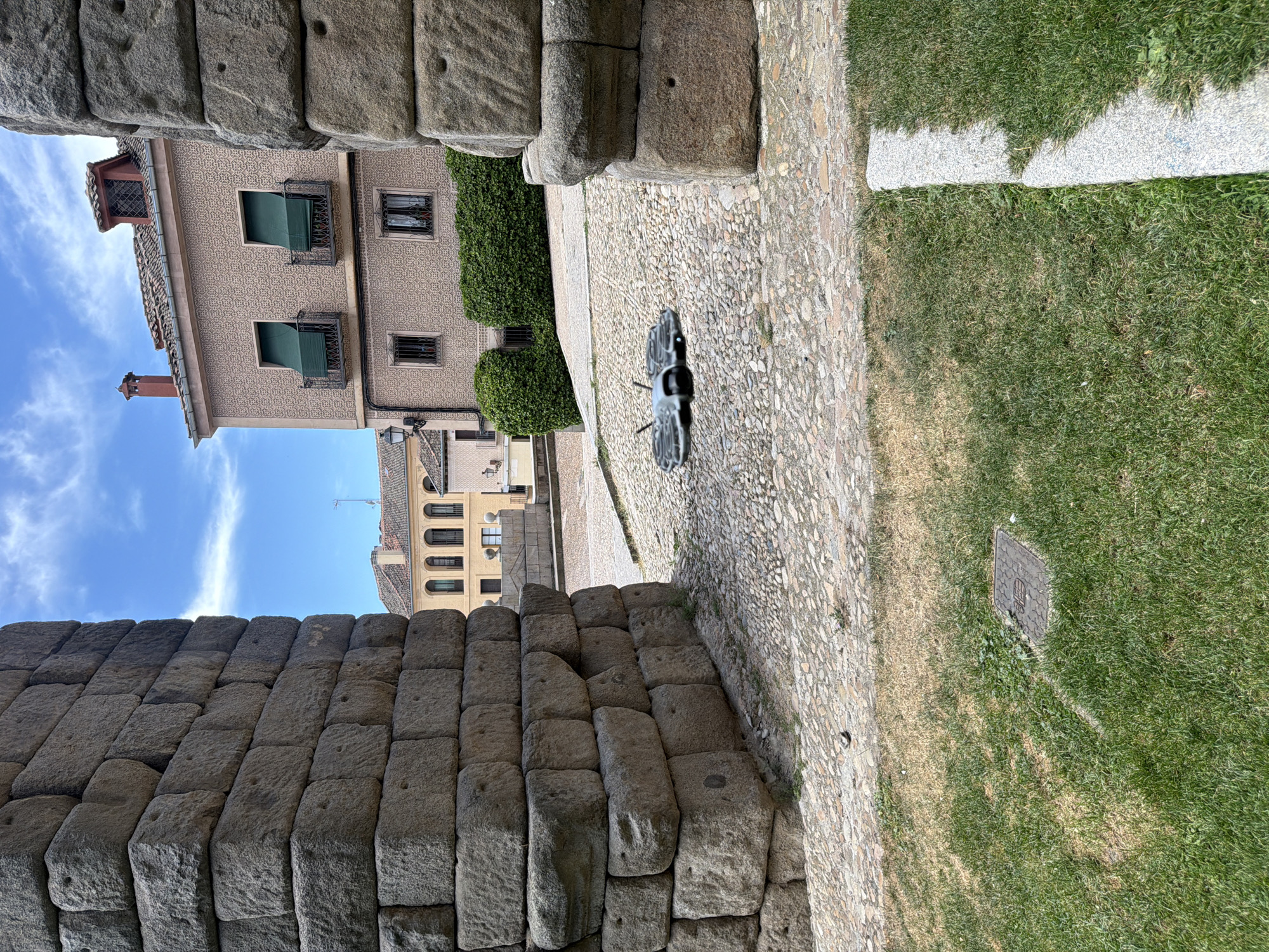 The image shows a drone flying low over a cobblestone plaza in Segovia, Spain, with the ancient Roman Aqueduct of Segovia prominently featured on the left and right sides of the frame. The aqueduct is constructed of massive granite blocks. In the background, a building with a pale pink facade, green awnings, and a terracotta tile roof is visible. The ground is a mix of grass and cobblestones. The sky is blue with scattered white clouds.