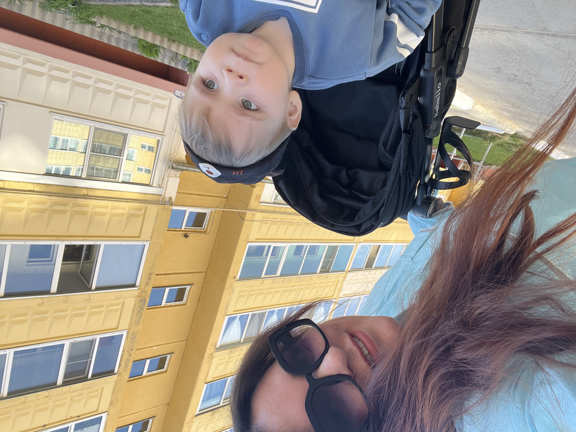 A selfie-style image shows a woman and a young child in a stroller, with an apartment building in the background. The woman is wearing sunglasses and has brown hair. The child, with blond hair and a hat, is in the stroller. The stroller is black. The apartment building is yellow. The location may be Noviy, Russia.