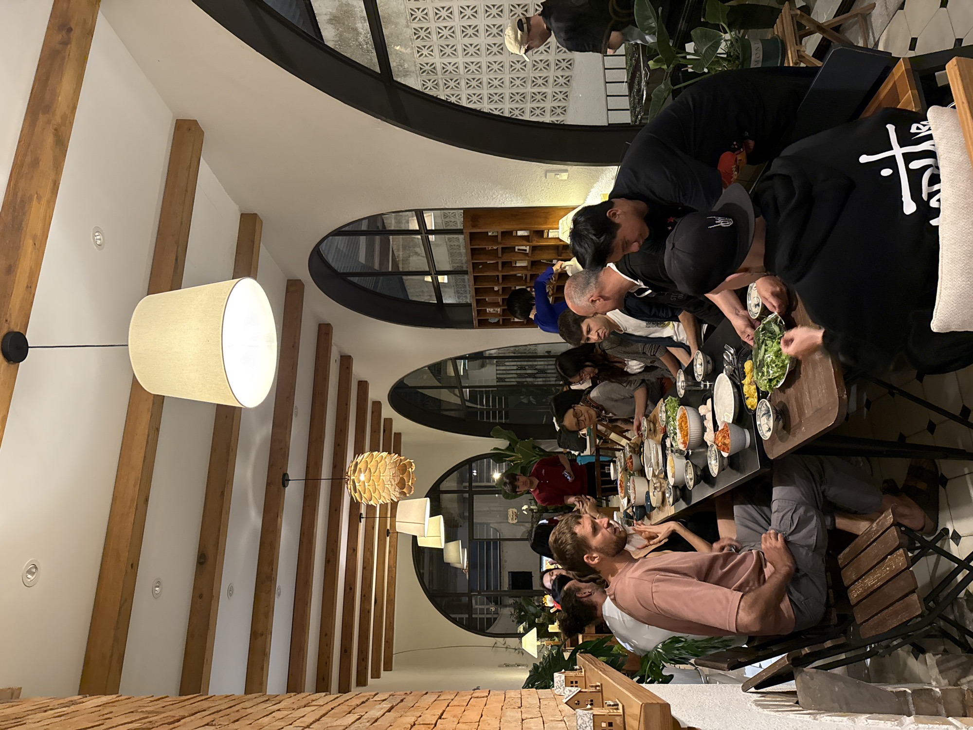 The image depicts a group of people gathered around a long dining table at a restaurant. The setting appears to be indoors, with a rustic yet modern aesthetic. Exposed wooden beams are visible on the ceiling, complementing the brick wall on the left side of the frame. Arched windows, some with decorative lattice work, are present in the background. Several people are seated at the table, engaged in eating and conversation. The table is laden with various dishes, suggesting a shared meal. In the foreground, a person wearing a pink t-shirt and gray pants is seated, while another person in a black hoodie is also prominent. The lighting suggests it is evening, with pendant lights and a lamp illuminating the space. The scene is likely a restaurant in Lam Vien - Da Lat, Vietnam, given the presence of lush greenery and the architectural style.
