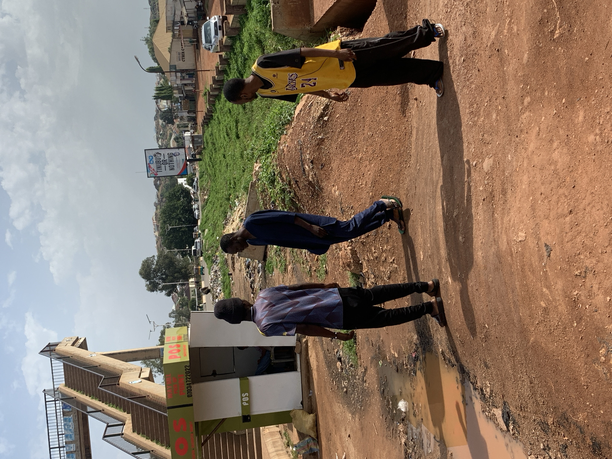 In Gura Top, Nigeria, three men are captured in a medium shot on a dirt path under a cloudy sky. The foreground shows a muddy puddle and reddish-brown dirt. In the mid-ground, the first man, seen from behind, wears a black beanie, a blue and purple patterned t-shirt, and black trousers with black sandals. To his right, a second man in a dark blue outfit with a matching head covering walks away, his body angled to the right. Further to the right, a third man faces left, wearing a yellow Los Angeles Lakers jersey with the number 24 and black trousers, standing near a concrete structure. Behind the men, a white kiosk labeled "POS" is visible, situated near a concrete pedestrian bridge with stairs and railings. In the background, there are buildings, green vegetation, roads with vehicles, and a billboard with the text "TINUBU -OR- NOTHING". The overall scene depicts an outdoor urban environment with signs of development and everyday life.