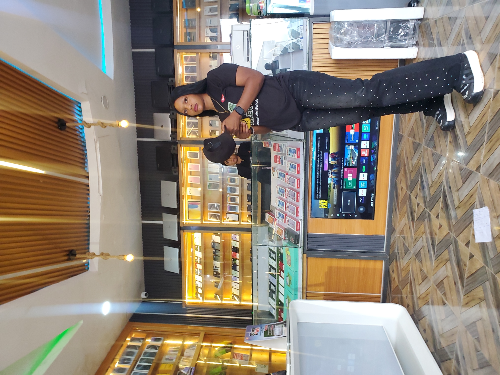 A full-body, eye-level shot captures a young Black woman standing in what appears to be a modern electronics store in Jos, Nigeria. She is wearing a black t-shirt with a graphic print, black jeans heavily adorned with small white studs, and platform sneakers. She holds a black baseball cap in her right hand, near her chest, and looks towards her left. Behind her, shelves are packed with mobile phones and other electronic devices, illuminated by warm lighting. A large television screen displays a menu of apps. The store's interior features wood-paneled ceilings and a patterned tiled floor. A younger woman, likely an employee, is partially visible in the background, standing behind a counter. The scene suggests a commercial environment focused on the sale of electronic goods.