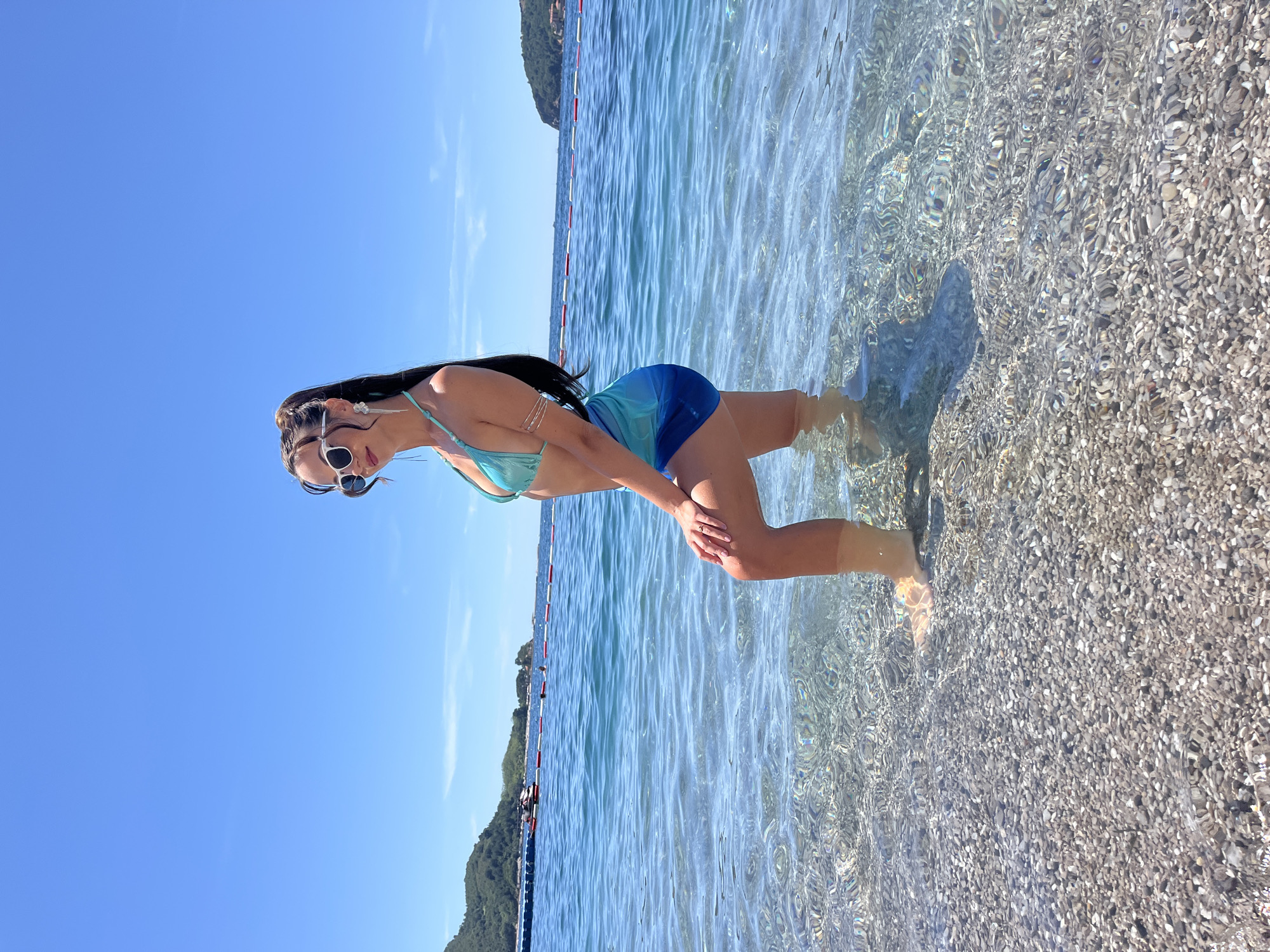 A woman stands in shallow, clear turquoise water near a pebbled beach, with the Mediterranean Sea stretching out behind her. She wears a turquoise bikini and a matching blue and green sarong. Her sunglasses have white frames, and she has long, dark hair pulled back in a ponytail, adorned with large, dangling earrings. She is posing with one knee bent and her hand resting on it. The water is calm with gentle ripples, and the sun is shining brightly, casting shimmering patterns on the seabed. In the background, a floating barrier marks off a swimming area, and distant green hills rise from the coastline, suggesting a view of Budva, Montenegro. The sky is a clear, vibrant blue.
