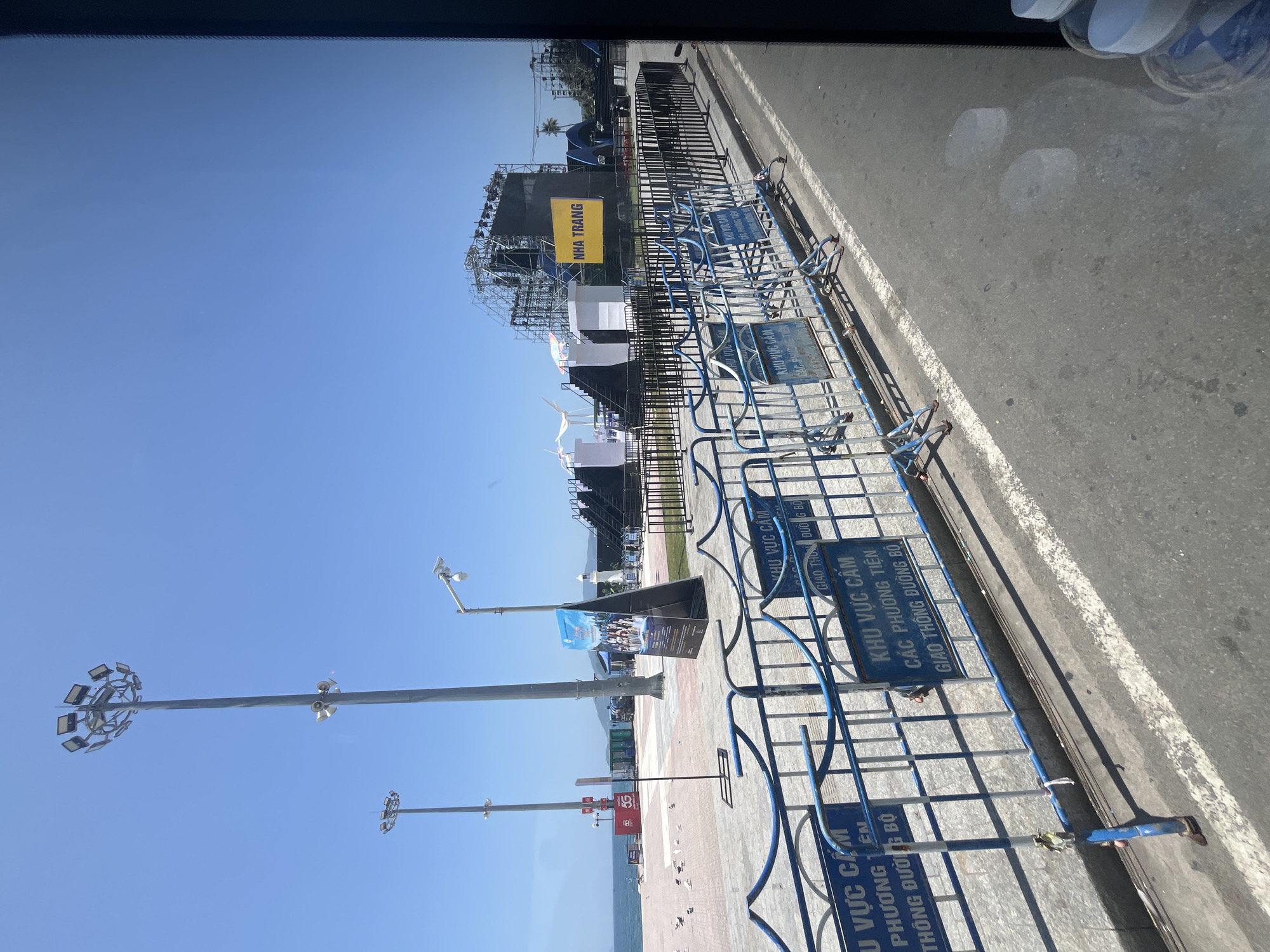 The image shows a view of a street in Nha Trang, Vietnam, with a clear blue sky and a body of water in the background. In the foreground, there are temporary metal barriers with blue trim, some of which have signs with Vietnamese text indicating a restricted area. Behind the barriers, there is a paved walkway with some scattered birds. Further back, there are tall light poles, a large stage setup with scaffolding and lighting equipment, and a yellow sign that reads "NHA TRANG". The scene appears to be a public area being prepared for an event.
