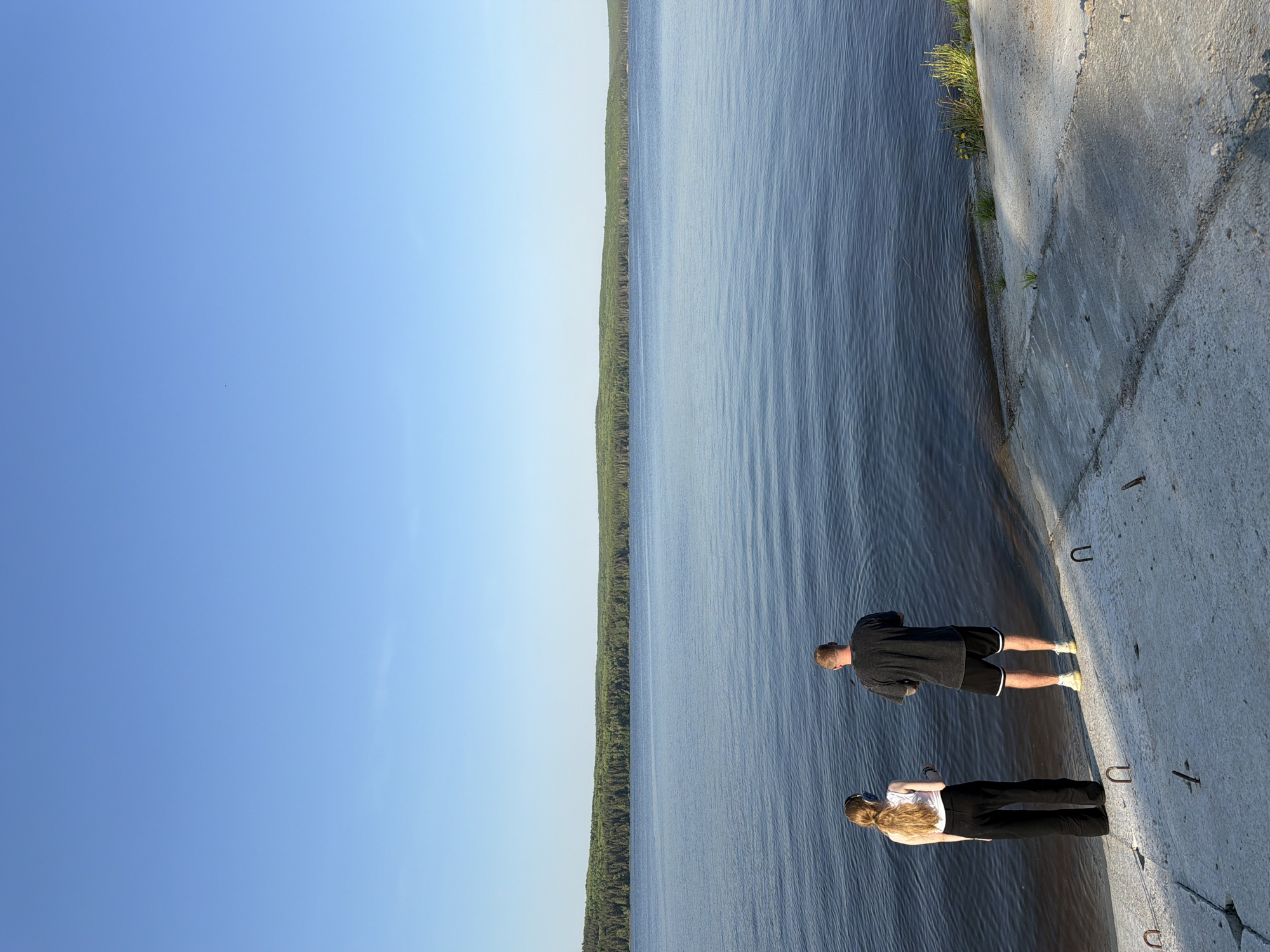 Two people, a man and a woman, stand on a concrete structure, possibly a dam or pier, next to a large body of water. A forest lines the opposite shore. The sky is clear and blue.
This scene likely takes place in or near the city of Chaikovsky, Russia, which is situated on the Kama River and has a dam, visible in the image.
