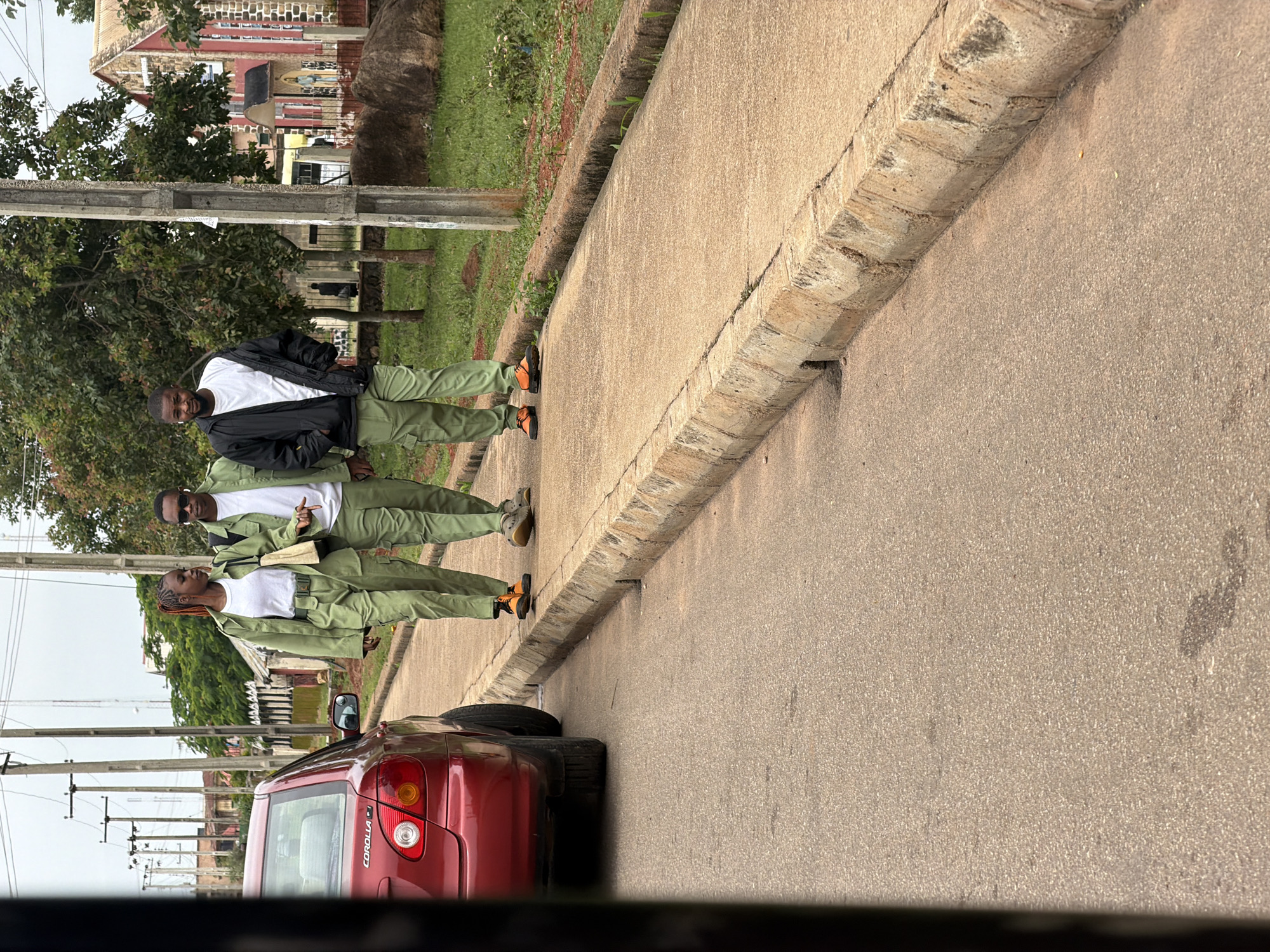 Three young adults, two men and one woman, are walking on a paved sidewalk next to a street in Bukuru, Nigeria. The woman and one of the men are wearing matching olive green uniforms consisting of a jacket and pants. The woman is wearing a white t-shirt underneath her jacket and carrying a tan tote bag. The man next to her is wearing a white t-shirt and sunglasses. The second man is wearing a black jacket over a white t-shirt and olive green pants. He is holding the zipper of his jacket. The three individuals are walking in the same direction on the sidewalk. To their left, a red Toyota Corolla is parked on the street. The environment includes trees, utility poles, and a building with a red facade in the background, suggesting an urban or suburban setting. The sky is overcast.