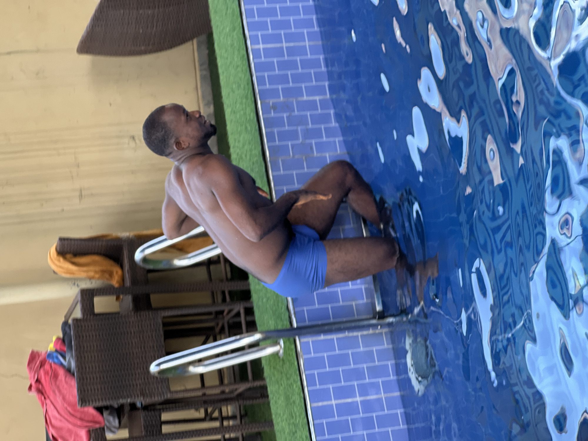 A shirtless man wearing blue swim trunks sits on the edge of a swimming pool with his legs submerged in the water. He is holding onto the pool's ladder and looking upwards. The pool is surrounded by blue tiles and green artificial turf. A wicker furniture set with clothing is visible in the background. The location is Unknown, Unknown.