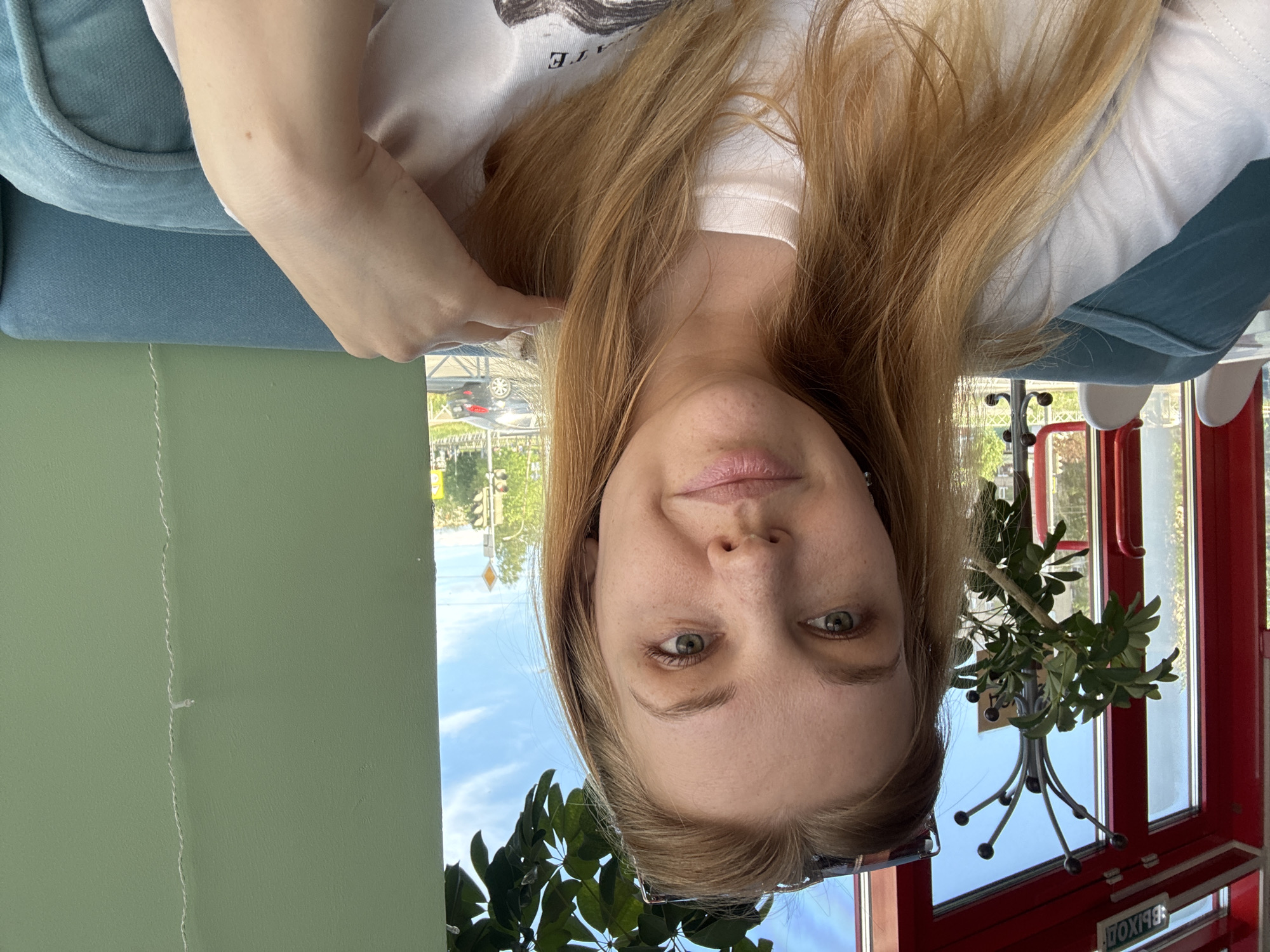 A woman with long, light brown hair is sitting in a cafe. She is wearing a white t-shirt. A green wall is visible to her left and a red window frame and potted plant are to her right. Outside the window, the sky is blue and there are buildings and trees. Given the location, the scene is likely within or near the Rusanovo district of Moscow, Russia.