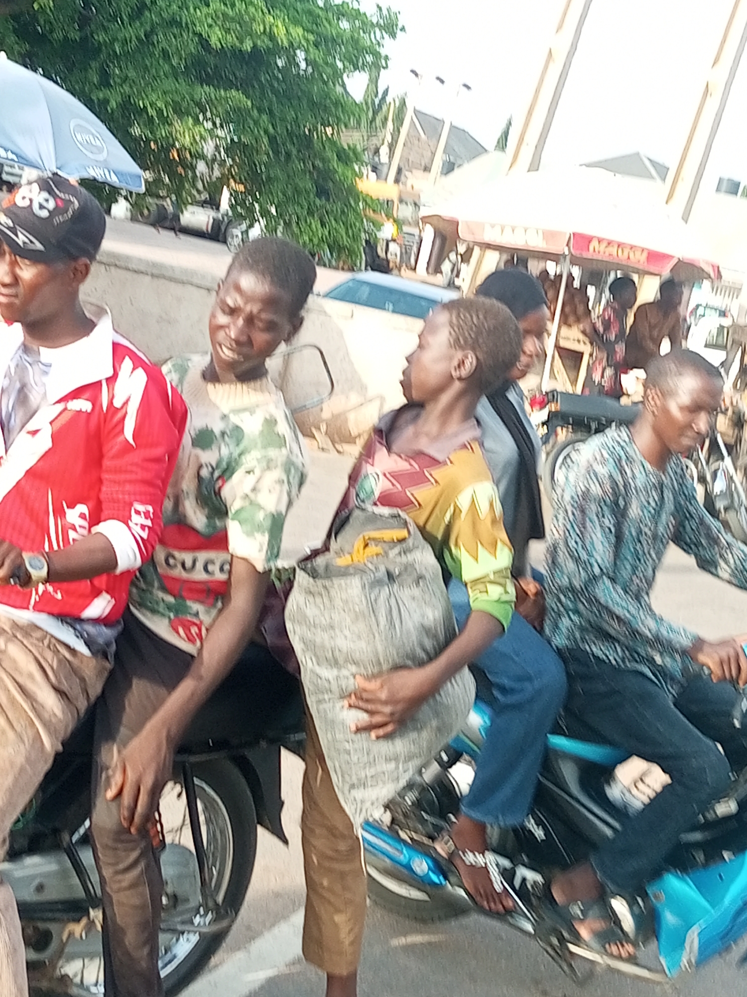 The image shows a group of people on a motorcycle in an outdoor setting, possibly a street or market. Four young men are prominently featured, with three of them riding on the motorcycle and one standing or walking beside it.
The person in the front left is wearing a red and white jacket and a black baseball cap. Next to him, on the motorcycle, is a person in a camouflage-patterned shirt with red accents, holding onto a large, light-colored bag. Behind them, another young man in a patterned shirt and blue jeans is also on the motorcycle. The person in the front right, who appears to be driving, is wearing a blue and white patterned long-sleeved shirt.
The scene is likely taking place during the daytime, given the bright lighting and shadows. There are trees and some sort of market stalls or awnings in the background, suggesting an urban or semi-urban environment. The overall mood seems casual, with the individuals appearing to be engaged in transit or a social gathering.
One person's shirt has the word "CUCO" visible on it. The blue motorcycle has a bright, possibly metallic finish. The general impression is of a vibrant, active scene with a focus on the people and their mode of transportation.