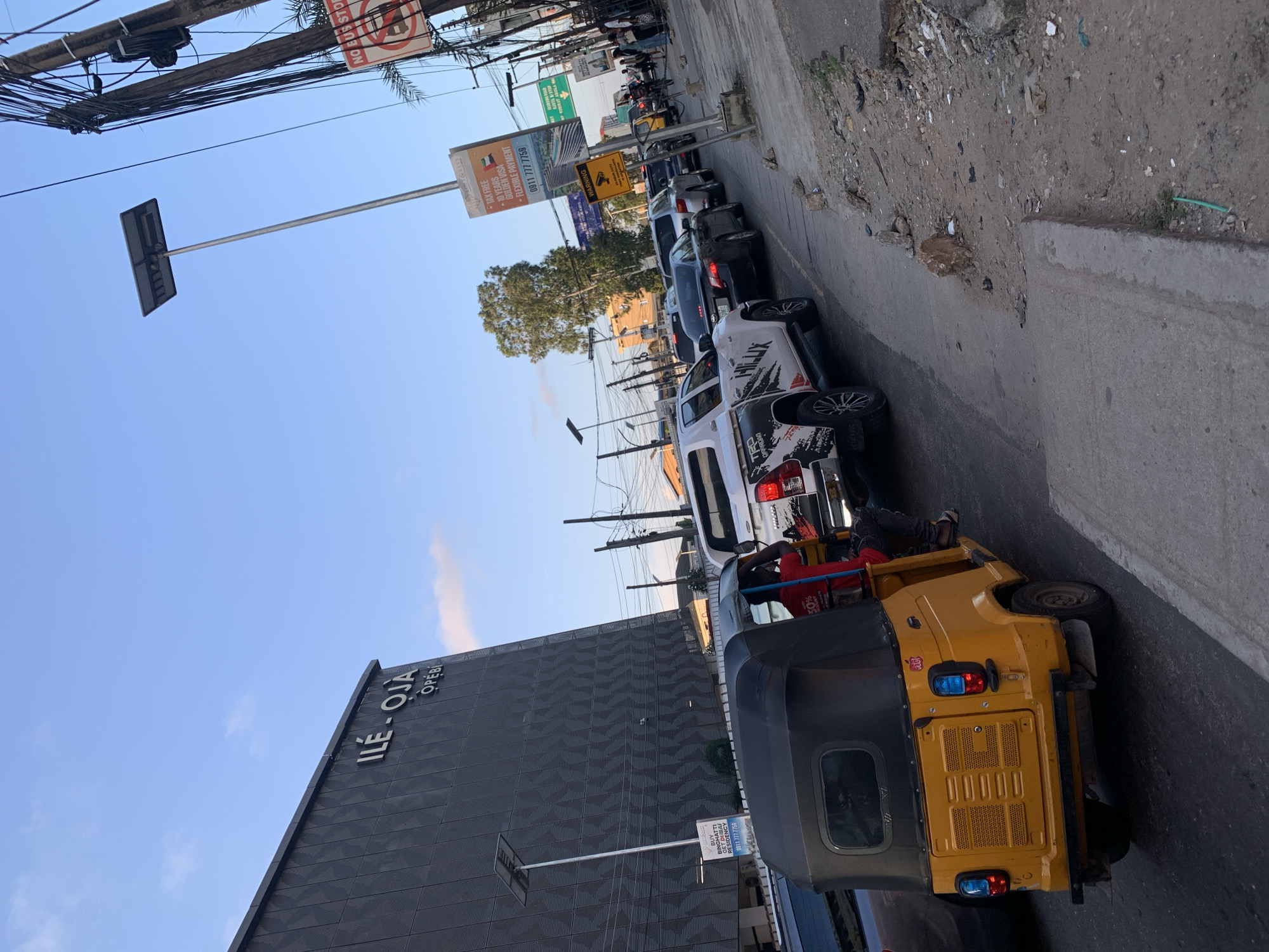 The image depicts a street scene in Ikeja, Nigeria, likely during a period of heavy traffic. In the foreground, a yellow auto-rickshaw, also known as a Keke Napep, is visible. The rickshaw has a black roof and is occupied by a driver wearing a red shirt and dark pants. The rickshaw is positioned on the right side of the road, facing away from the camera.
Behind the auto-rickshaw, a line of vehicles is backed up, indicating traffic congestion. Among these vehicles is a white pickup truck with "HILUX" branding and a distinctive graphic design on its side. Other cars, including a black sedan, are also present in the traffic jam.
To the left of the street, a modern, dark gray building with the signage "ILÉ-ỌJÀ OPEBI" is prominently featured. The building has a textured facade and appears to be an office or commercial structure. A solar-powered street light stands in front of the building.
Further down the street, more traffic and a variety of signs are visible. One sign reads "NO BUS STOP" with a red circle and a diagonal line through a bus symbol. Another tall sign displays advertisements, including contact information and images of buildings. Overhead, a complex network of utility poles and electrical wires crisscrosses the sky, a common sight in many urban environments. The sky is blue with a few scattered clouds, suggesting it is either early morning or late afternoon. The ground on the side of the road is unpaved and littered with debris, typical of urban infrastructure in some areas.