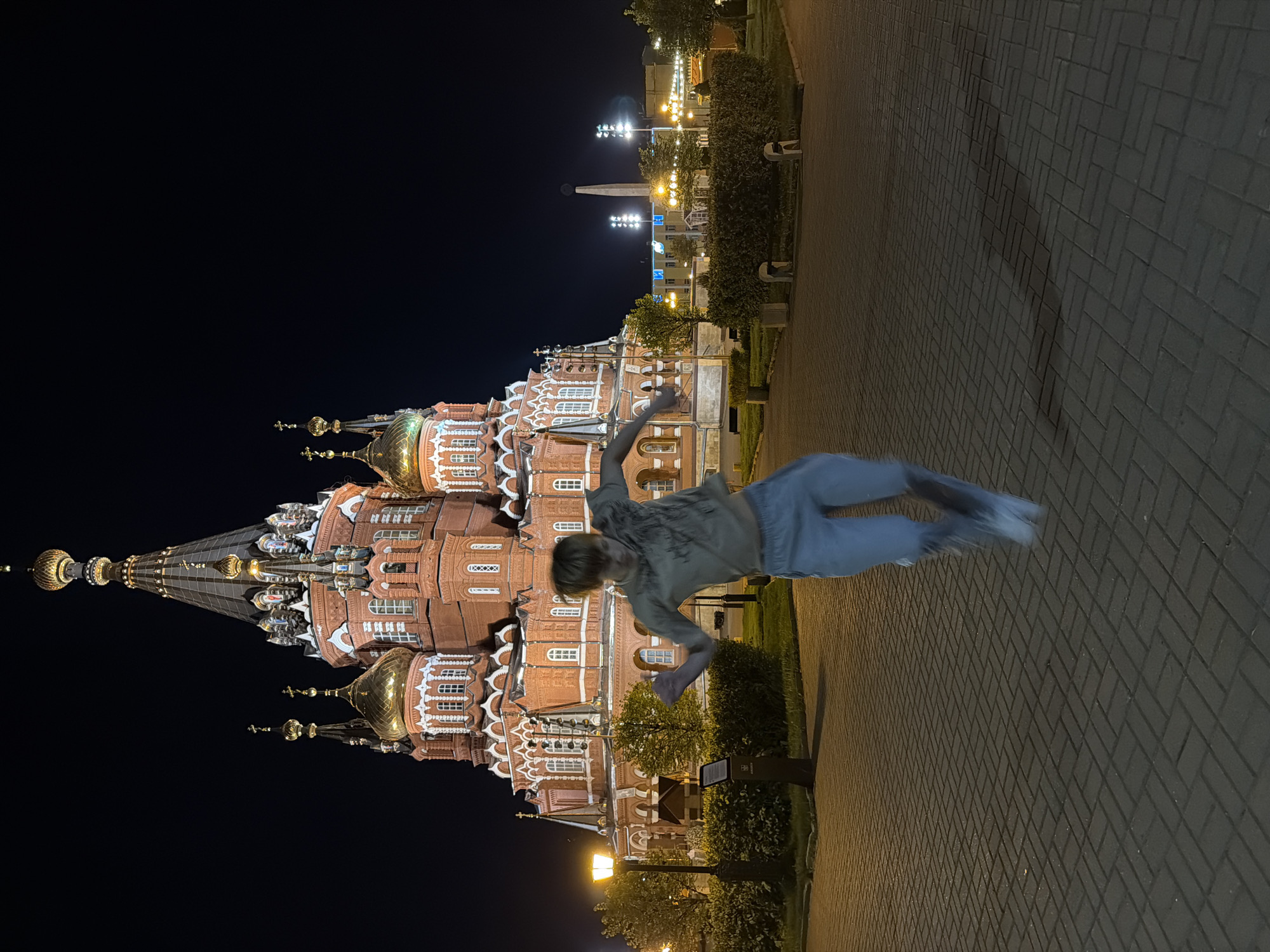 The image shows a person in mid-air, seemingly performing a gymnastic move, in front of St. Michael's Cathedral in Izhevsk, Russia, at night. The cathedral is illuminated, showcasing its onion domes and intricate brickwork. The sky is dark, and the foreground consists of a brick walkway and some greenery. Streetlights and other buildings can be seen in the background.