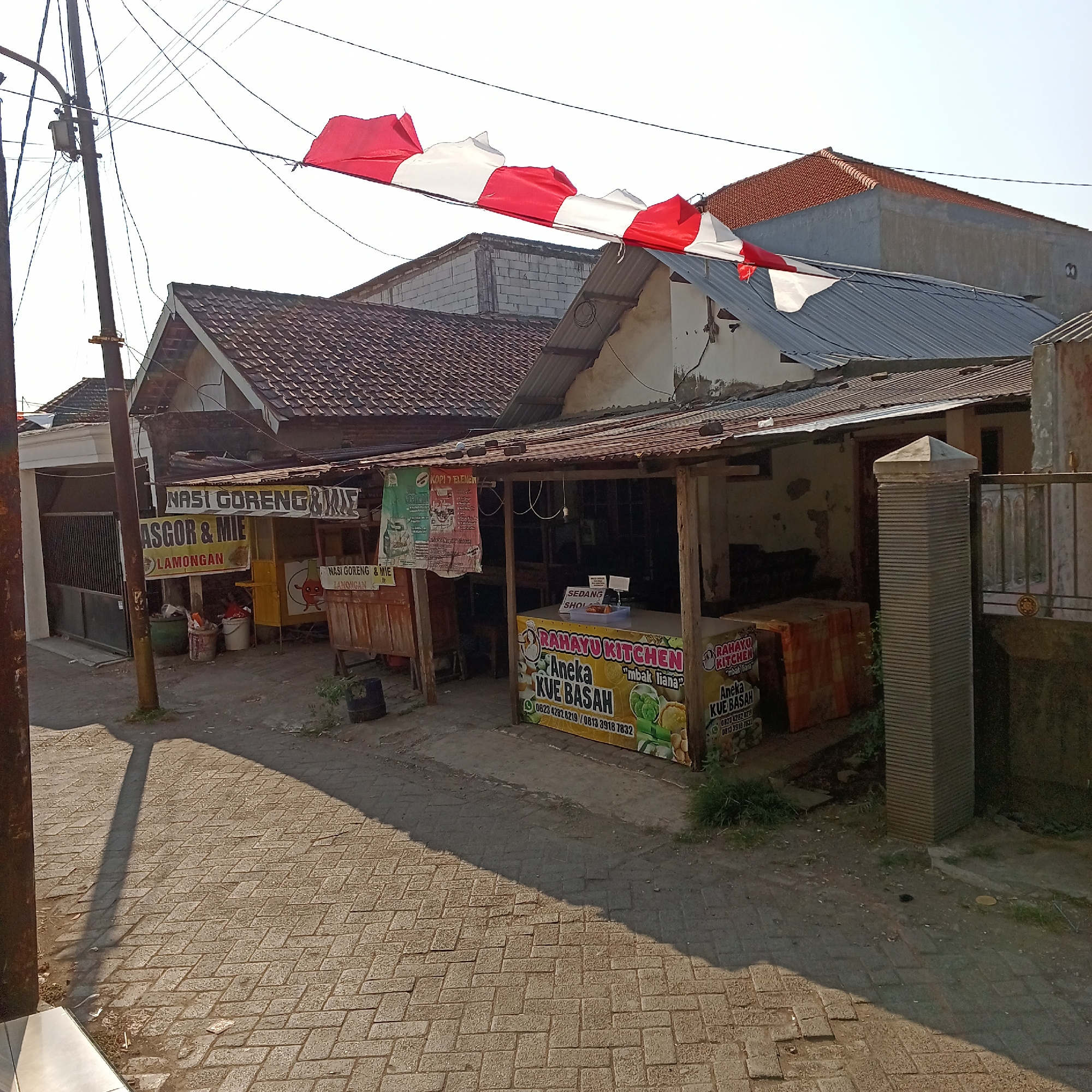 The image captures a street scene in Kepuh Kiriman, Indonesia, featuring a row of food stalls and residential buildings under a bright sky. In the foreground, a paved walkway with a diamond pattern is visible, casting long shadows from overhead utility poles and wires. A red and white Indonesian flag is stretched across wires in the upper portion of the frame, appearing to be a celebratory decoration.
To the left, a dark metal gate leads to a property with a white facade. Adjacent to this gate, a utility pole stands tall, adorned with electrical transformers and a multitude of wires. Further along the street, a food stall is visible with a sign that reads "NASI GORENG & MIE ASGOR & MIE LAMONGAN." Several buckets and containers are placed near this stall.
The central focus of the image is a food stall with a corrugated metal roof, displaying a banner that advertises "RAHAYU KITCHEN Aneka KUE BASAH" (Various Wet Cakes) with contact information. This stall appears to be actively operating, with some items displayed on its counter. Another sign, also for "NASI GORENG & MIE LAMONGAN," is partially visible on a wooden structure to the right of the primary stall.
Behind the stalls, residential buildings with red-tiled roofs and varying architectural styles are present. The overall environment suggests a typical Indonesian street scene with local businesses and everyday life. The lighting indicates it is likely daytime, with the sun creating distinct shadows on the ground.