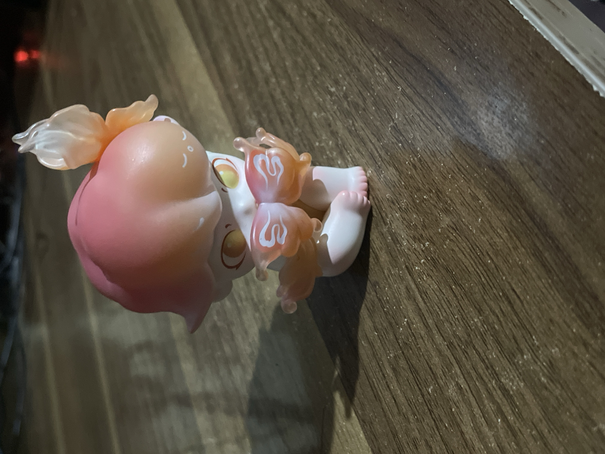 A small, stylized toy figure is depicted lying on its back on a dark wooden surface. The figure, which appears to be a cherubic character or a baby, has a soft, rounded form. Its head and hair feature a gradient of pink and orange, complementing its pale white body. Translucent, shell-like or petal-like adornments, with distinct white swirling patterns, are visible on the back of its head and around its wrists. The toy's face has large, dark, stylized eyes, and its small, plump feet with individual toes are visible.
The wooden surface beneath the toy is dark brown, exhibiting a clear wood grain pattern and scattered specks of white, possibly dust or small debris. The background is out of focus and dark, with a faint red light source visible in the distance on the left side of the frame. The scene suggests an indoor setting with low, artificial lighting, possibly in the evening. The lighting casts soft shadows from the toy onto the wooden surface. There is no visible text in the image.