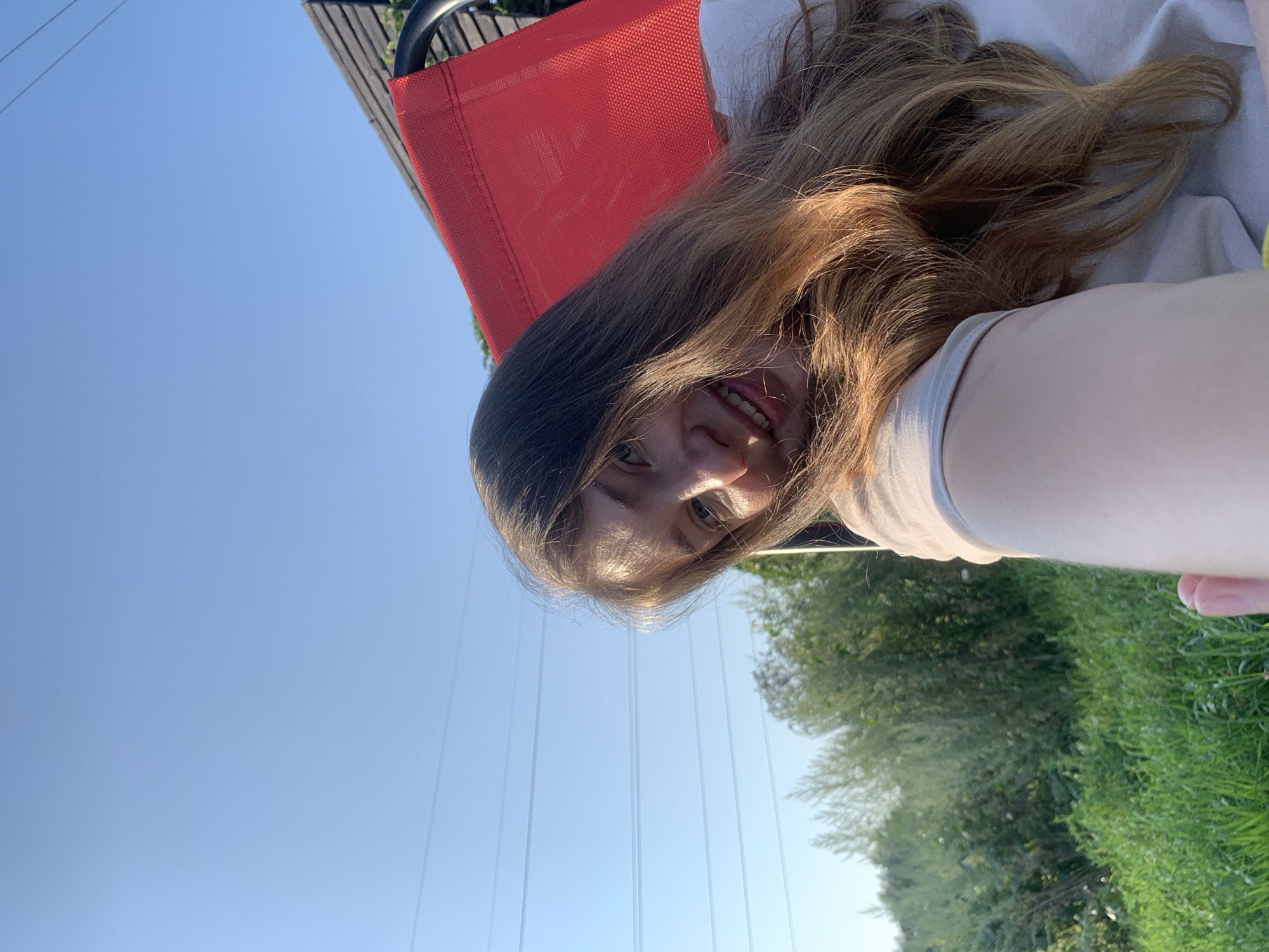 The image depicts a young woman in a selfie, taken outdoors on a clear day. She is seated in a red chair, with her head tilted back and hair falling over her shoulder. The background shows lush green grass and trees, suggesting a natural setting within the city of Chaikovsky, Russia. Power lines are visible in the distance, spanning across the bright blue sky. The woman is smiling and looking towards the camera, with sunlight illuminating her face and hair.