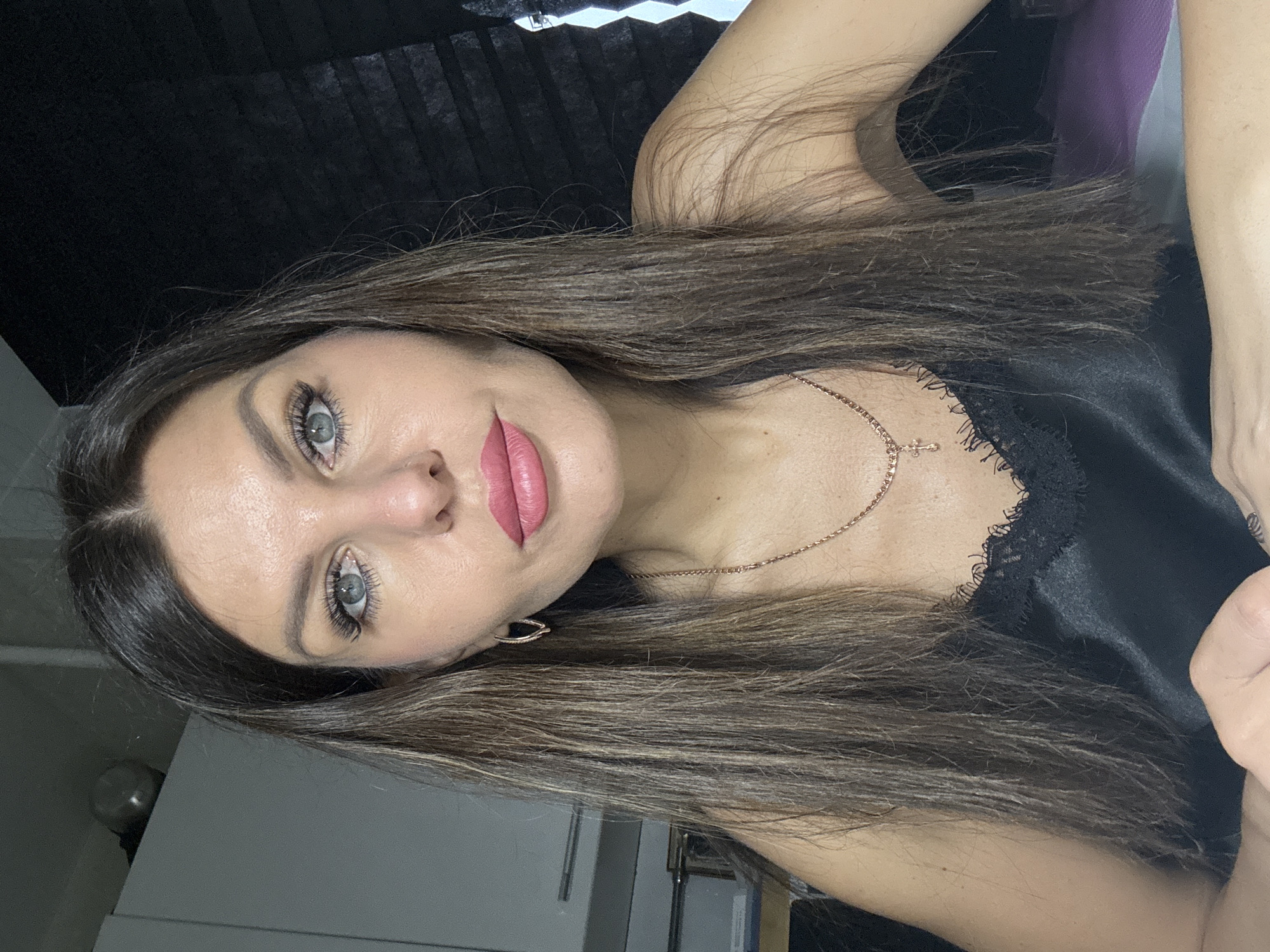 A woman with long, brown, highlighted hair, light blue eyes, and pink lipstick is visible. She is wearing a black satin top with lace trim and a gold necklace with a cross pendant. She appears to be indoors, based on the lighting and the presence of what seems to be kitchen cabinetry in the background. No evidence suggests this image was taken in Chaikovsky, Russia.