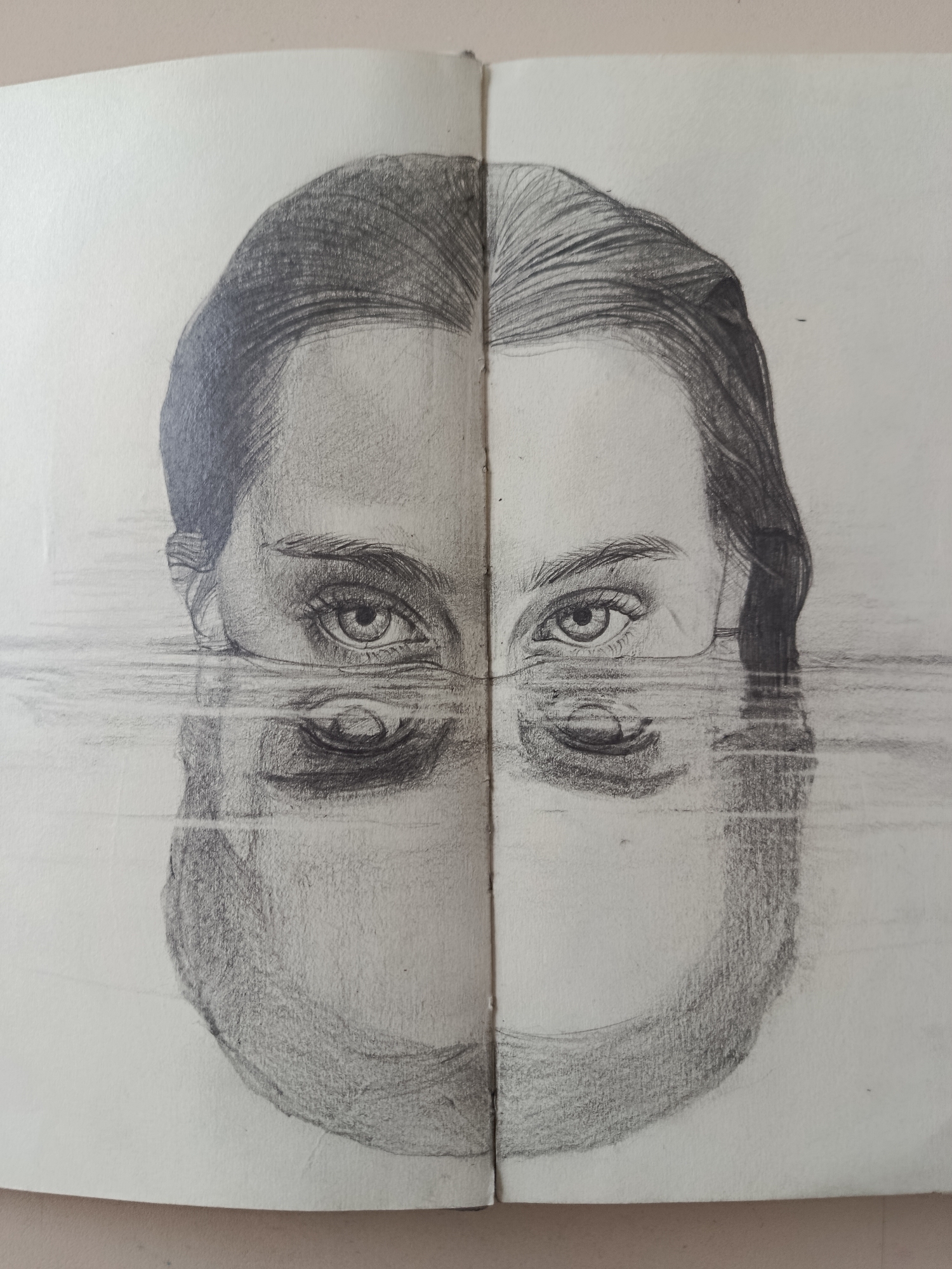 The media file presents a detailed pencil drawing of a human face, likely female, rendered across two pages of a sketchbook. The central fold of the sketchbook bisects the face vertically. The top portion of the face, including the forehead, eyes, and eyebrows, appears above a horizontal water line. The eyes are open and gaze directly forward, conveying a calm or pensive expression. Below the water line, the lower half of the face – the nose, mouth, and chin – is depicted as a reflection, heavily distorted and fragmented by ripples on the water's surface. These ripples are represented by prominent horizontal lines that span across both pages.
The drawing showcases a realistic artistic style with careful attention to shading and texture, particularly in the rendering of the hair, eyes, and the subtle contours of the face. Various pencil tones create depth and contrast. The hair is drawn with visible strokes, suggesting strands and movement. The texture of the paper is visible, adding to the handmade quality of the artwork. No specific location cues, time of day, or weather elements are explicitly depicted beyond the presence of water. There is no visible text in the image. The overall composition creates a surreal and introspective mood, highlighting themes of self-reflection or duality.