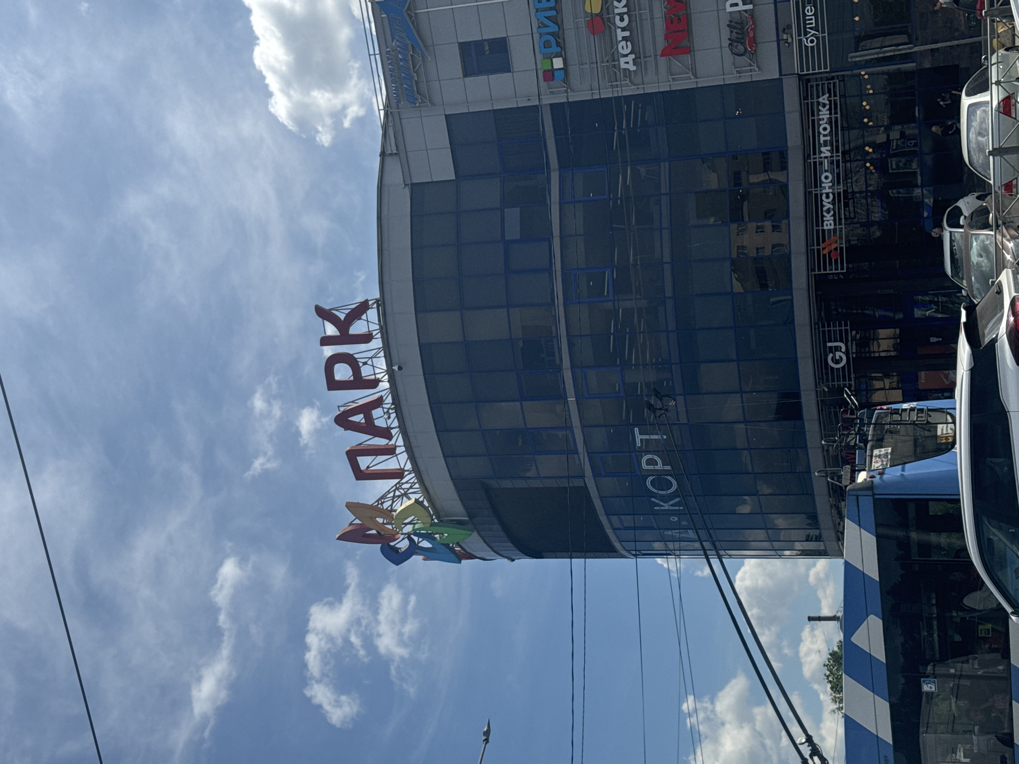 The image shows the exterior of a modern, multi-story building in Saint Petersburg, Russia. The building has a curved facade with large, dark blue tinted windows. A large, colorful sign at the top of the building reads "ПАРК" in red letters, with a stylized colorful pinwheel logo to its left. Below this, on the side of the building, are various signs for businesses, including "К-КОРТ," "РИВ," "детск," "NEW," "Chili P," and "буше." At street level, the sign "ВКУСНО — И ТОЧКА" is visible, a prominent fast-food chain. The foreground shows a blue and white striped public transport vehicle, possibly a trolleybus, and several cars parked or driving on the street. The sky above is blue with scattered white clouds, and power lines are visible across the top of the image. The overall scene depicts a busy urban environment.