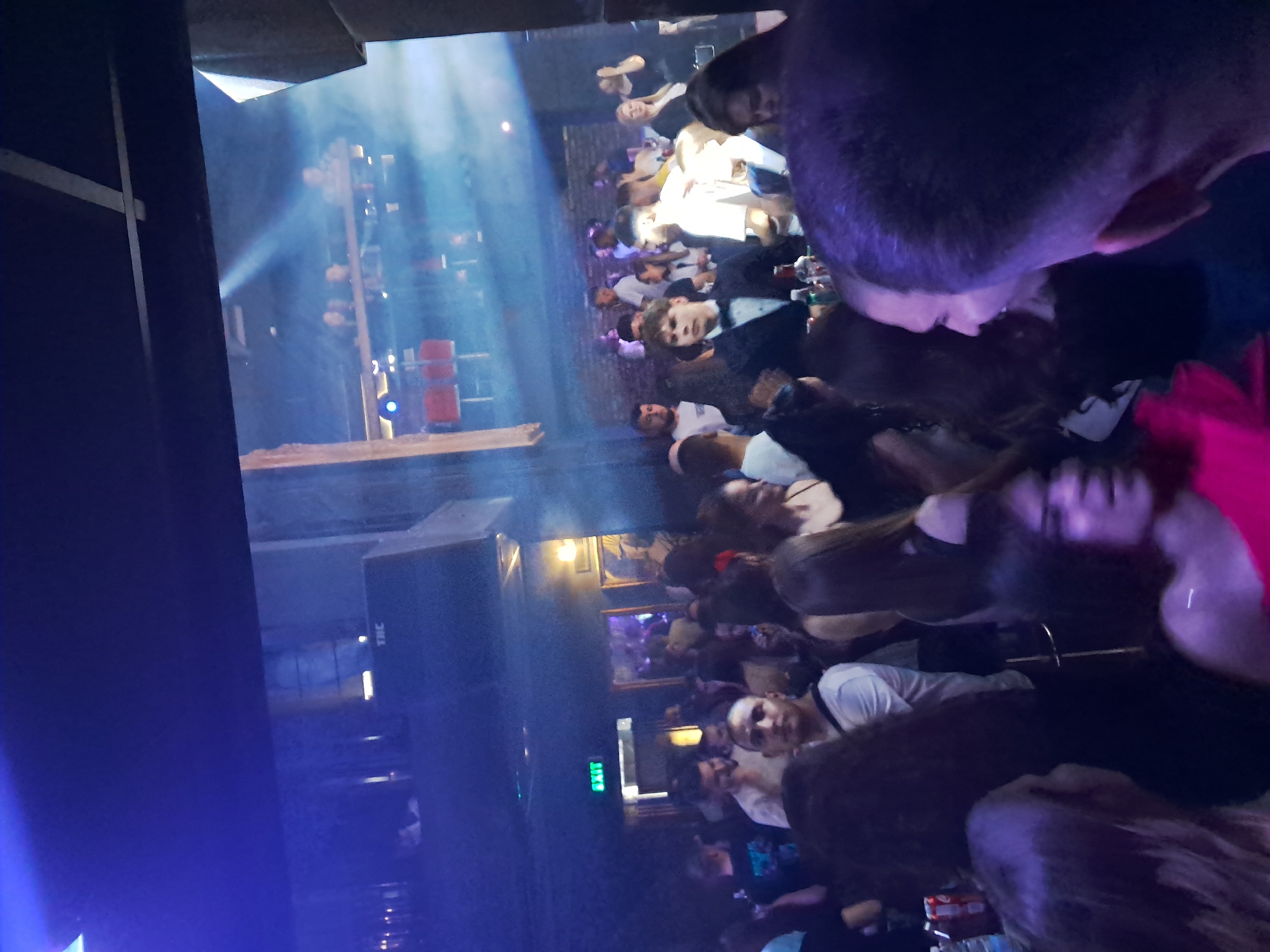 The image captures a crowded indoor social event, likely a club night or party, illuminated by atmospheric blue and purple lighting. The scene suggests it is nighttime in a venue located in Veliko Tarnovo, Bulgaria.
Numerous people, appearing to be young adults, are gathered throughout the space. Many are standing and socializing in groups, while others are seated at tables or bar areas. Several individuals are visible looking towards the camera, while others are engaged in conversation or observing their surroundings. Their expressions generally suggest engagement and enjoyment.
The environment features dim lighting with strong, distinct beams of light cutting through visible haze or smoke, contributing to a dynamic atmosphere. Visible elements include a wooden bar or railing, walls that appear to be brick, and framed pictures hanging on a back wall. Various drinks, including bottles and glasses, are seen on tables and surfaces. Large speakers or sound equipment are present on the left side of the frame.
A bright green, illuminated "EXIT" sign is clearly visible on a wall in the mid-ground on the left. The letters "THC" can also be faintly discerned on the side of a speaker or equipment. The overall impression is a lively social gathering taking place within a distinct club or event setting.